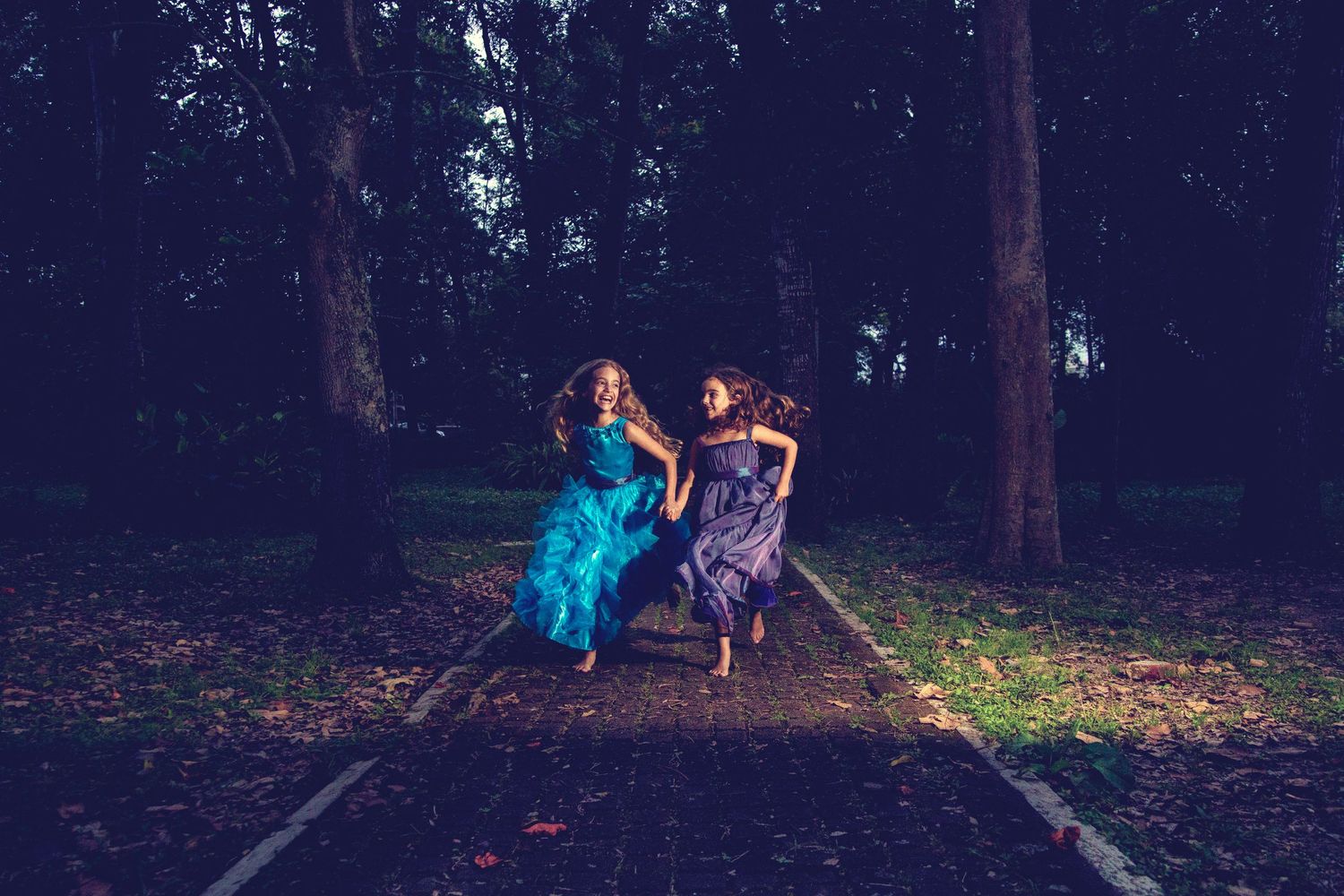 Two girls in colorful gowns run joyfully through a dark, moody forest path at dusk.