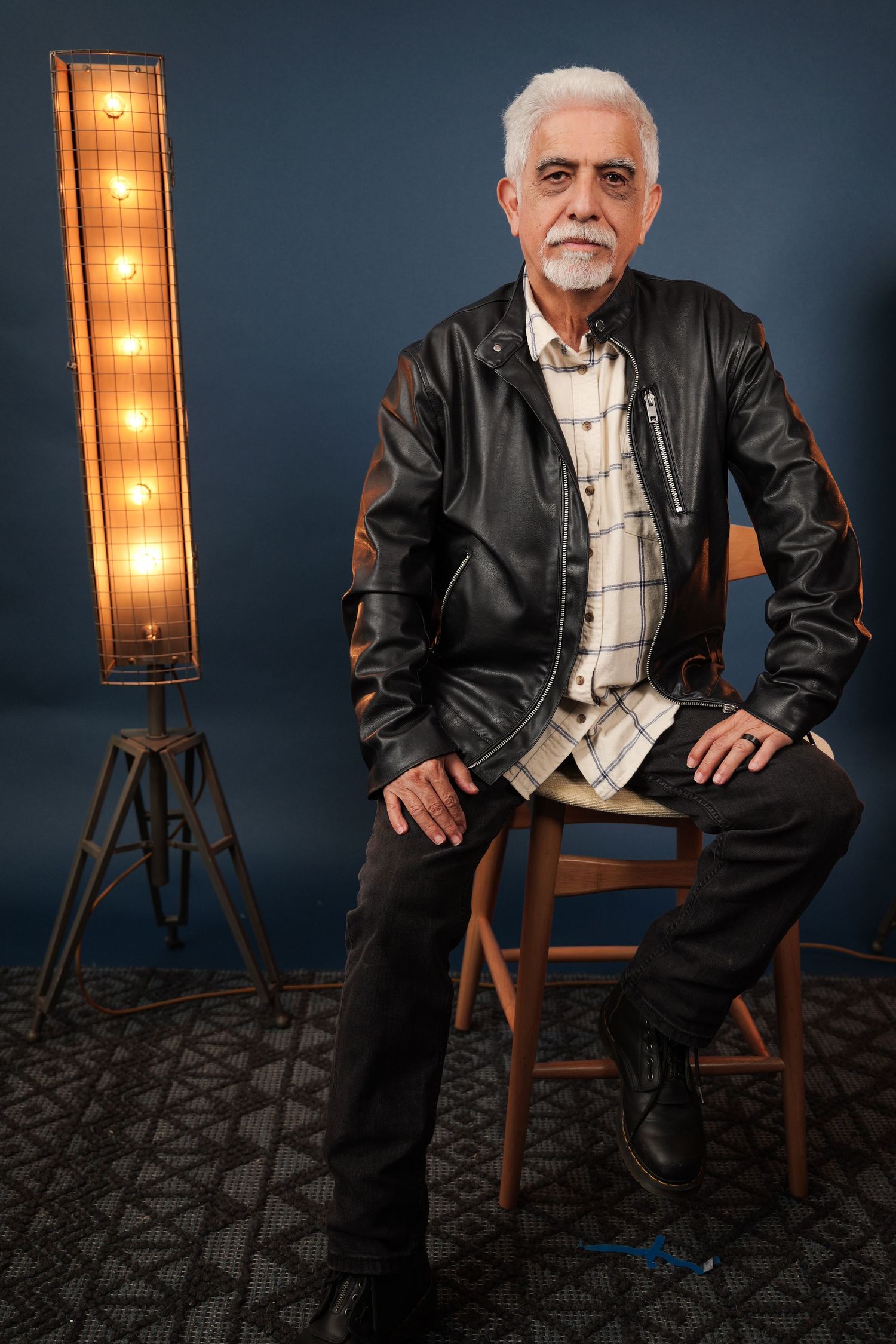 Older man with white hair in black leather jacket sits on wooden stool beside vintage floor lamp on dark blue background.