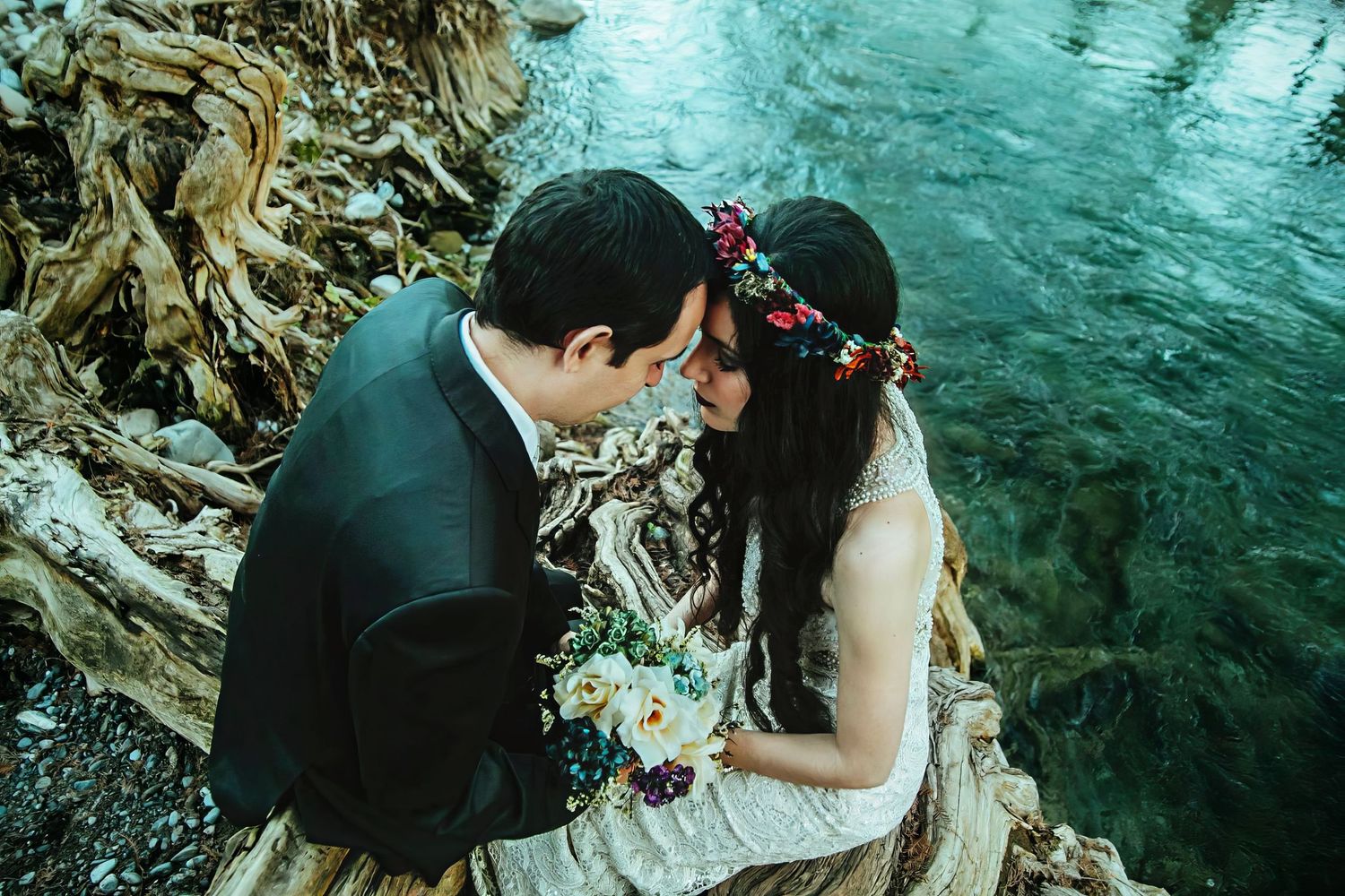 Bride and groom sharing a romantic moment by a river, bride wearing a floral crown and holding a bouquet.