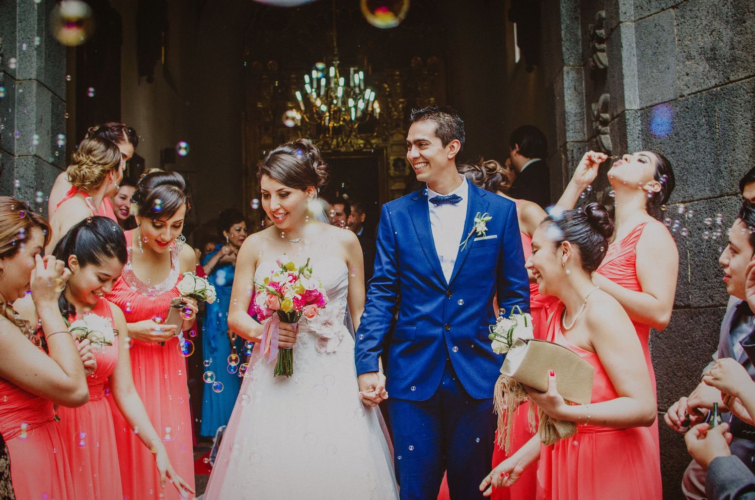 Bride and groom exit church ceremony surrounded by coral-dressed bridesmaids blowing bubbles in celebration.