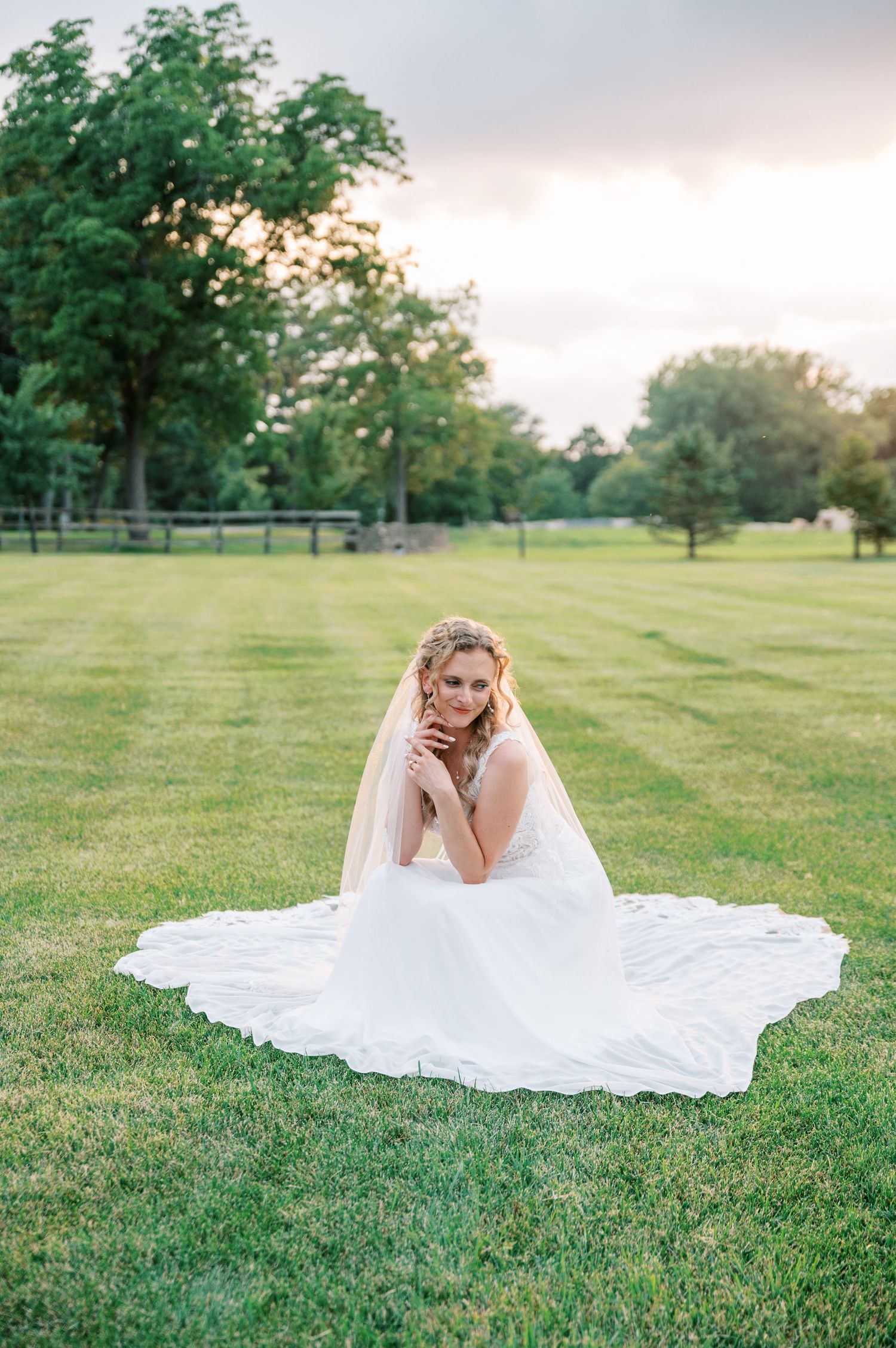 Bride in a flowing white gown and veil sits on a manicured green lawn with trees in the background at sunset.