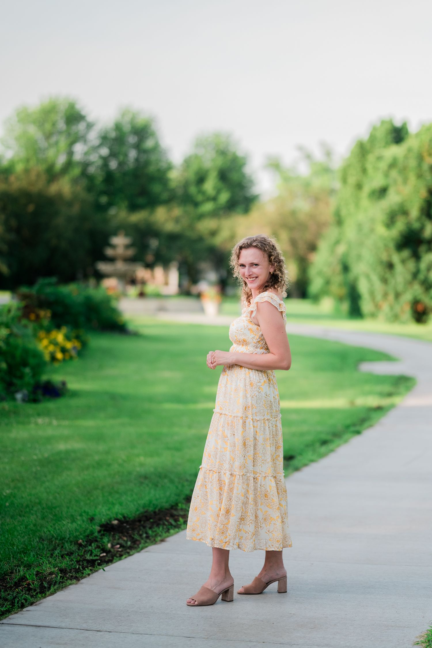 Woman with curly hair smiling in a yellow floral tiered sundress and tan heeled sandals on a sidewalk beside a green lawn and trees.