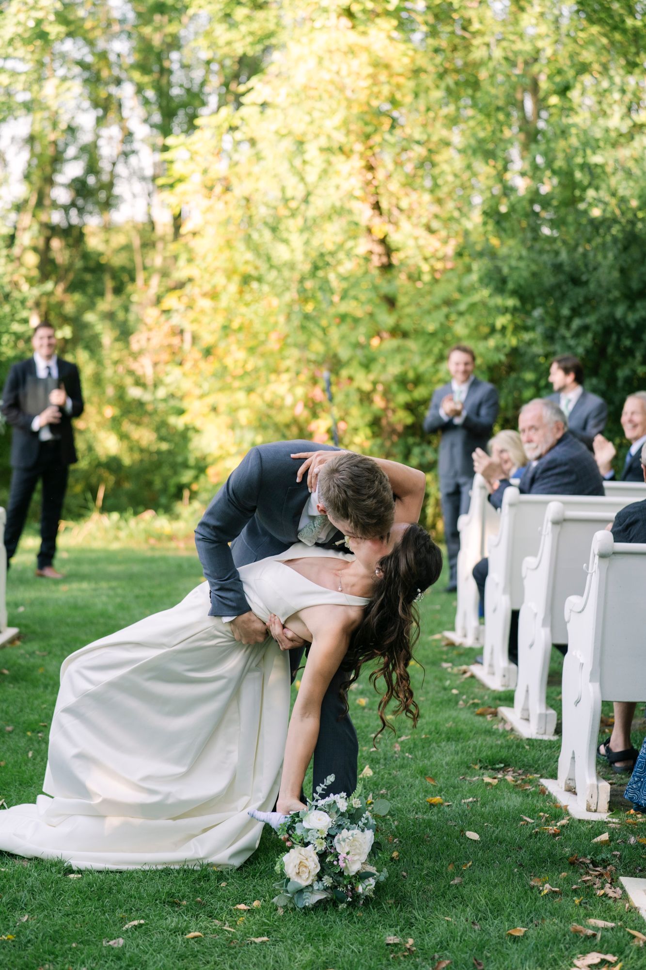 Groom dips and kisses his bride on an outdoor grass aisle as smiling wedding guests applaud from white wooden pews among autumn trees.