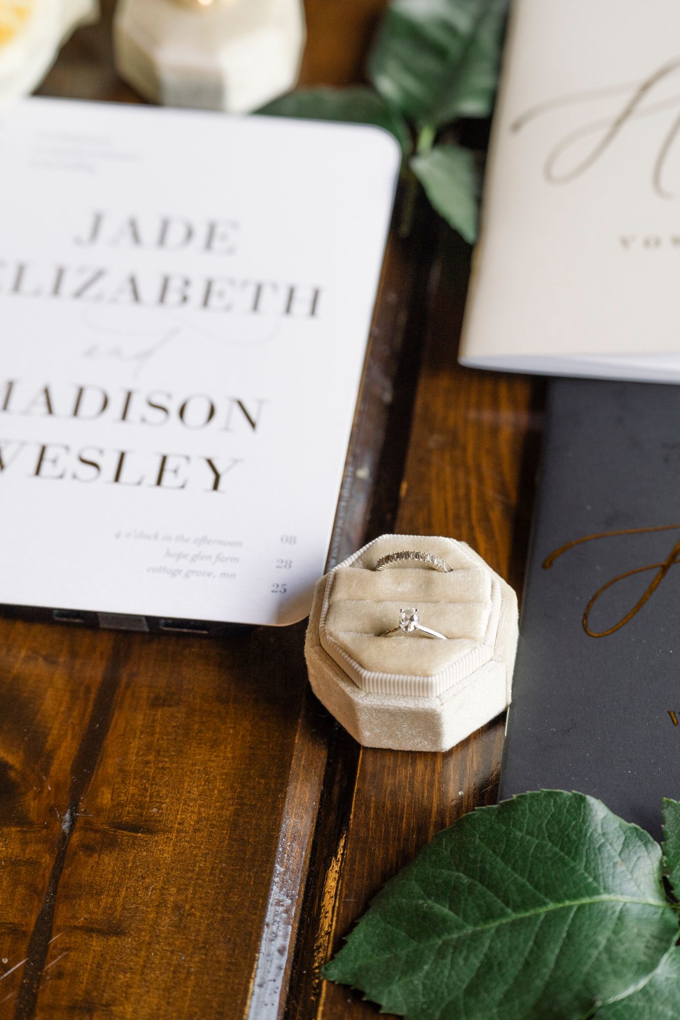 Engagement ring in a cream velvet octagonal box on a rustic wood table beside a wedding invitation, greenery, and gold-script stationery.