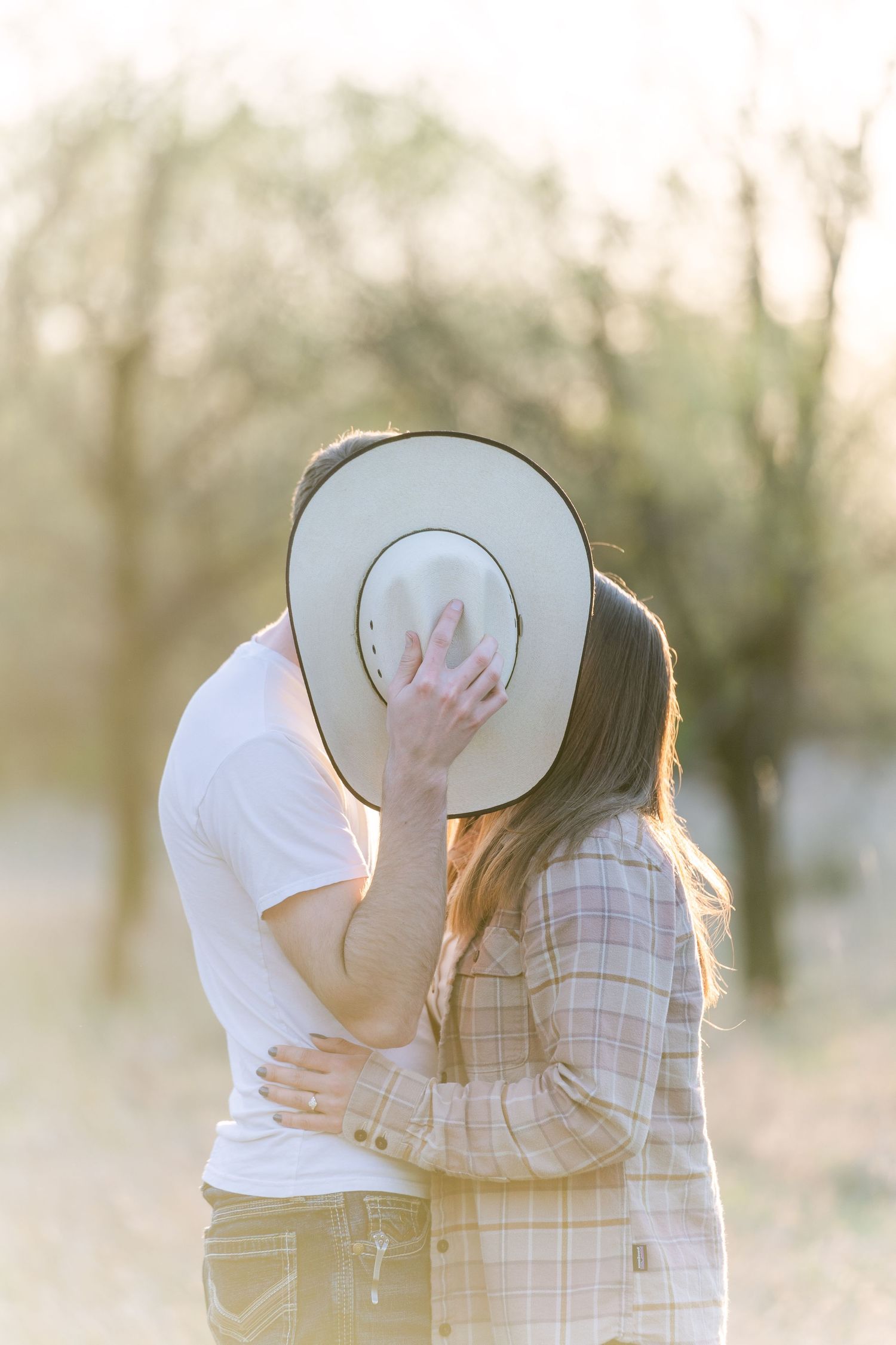 Couple in an orchard at golden hour kissing behind a white cowboy hat, he in a white tee, she in a plaid flannel.