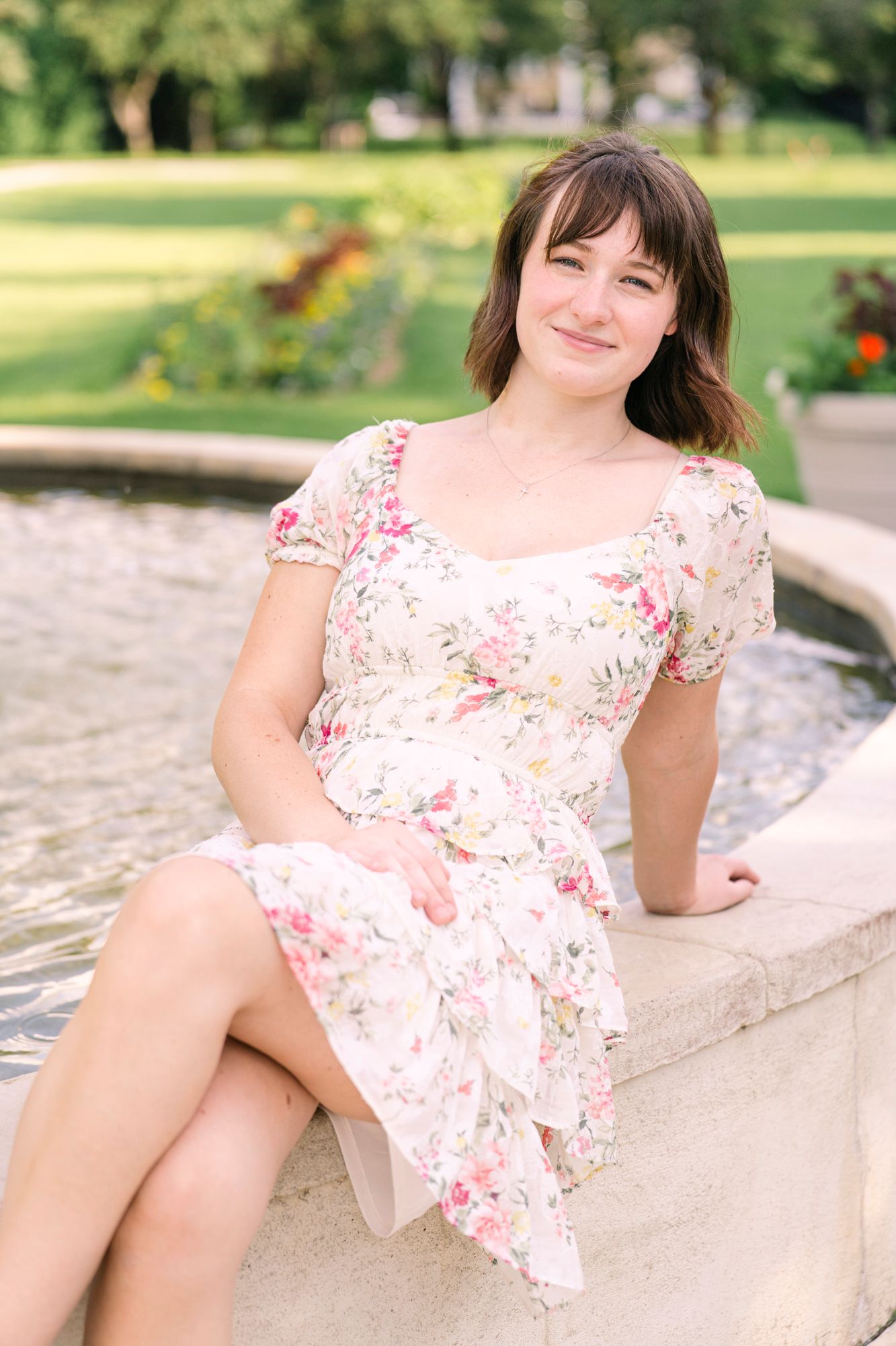 Floral and sunlit portrait of a high school-aged girl sitting by a fountain at Arneson Acres in Edina, Minnesota, smiling brightly at the ca