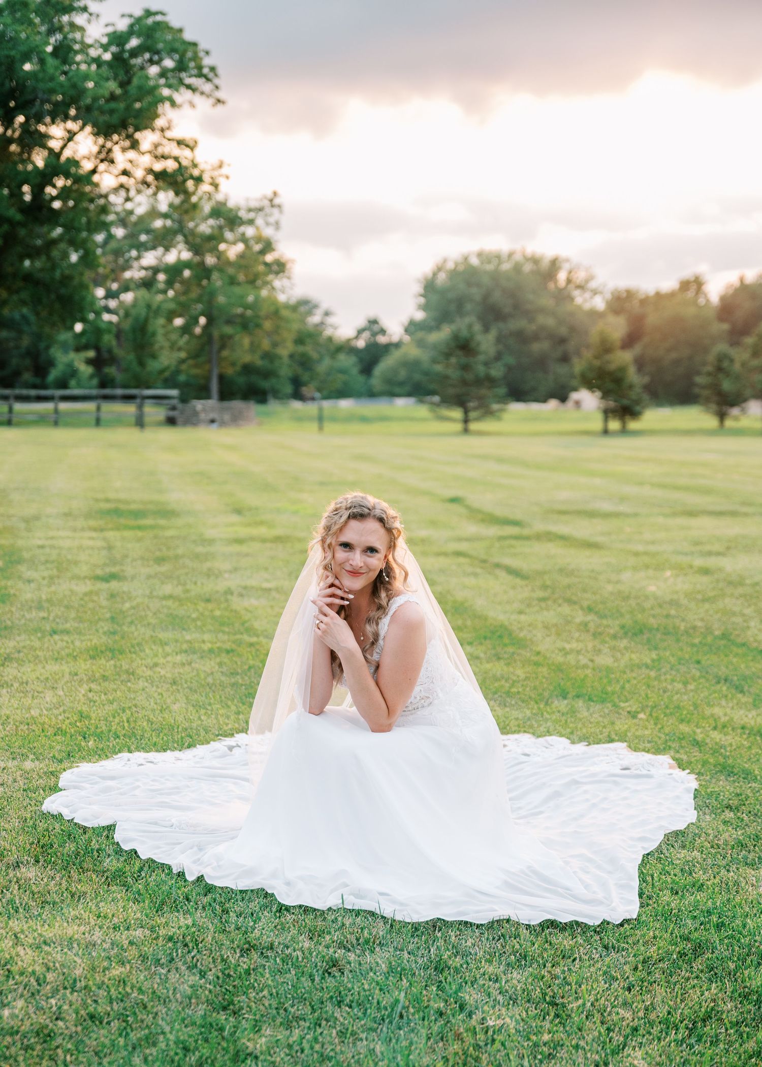 Smiling bride in a flowing white lace wedding gown and veil sits on a lush green lawn at sunset with trees in the background.