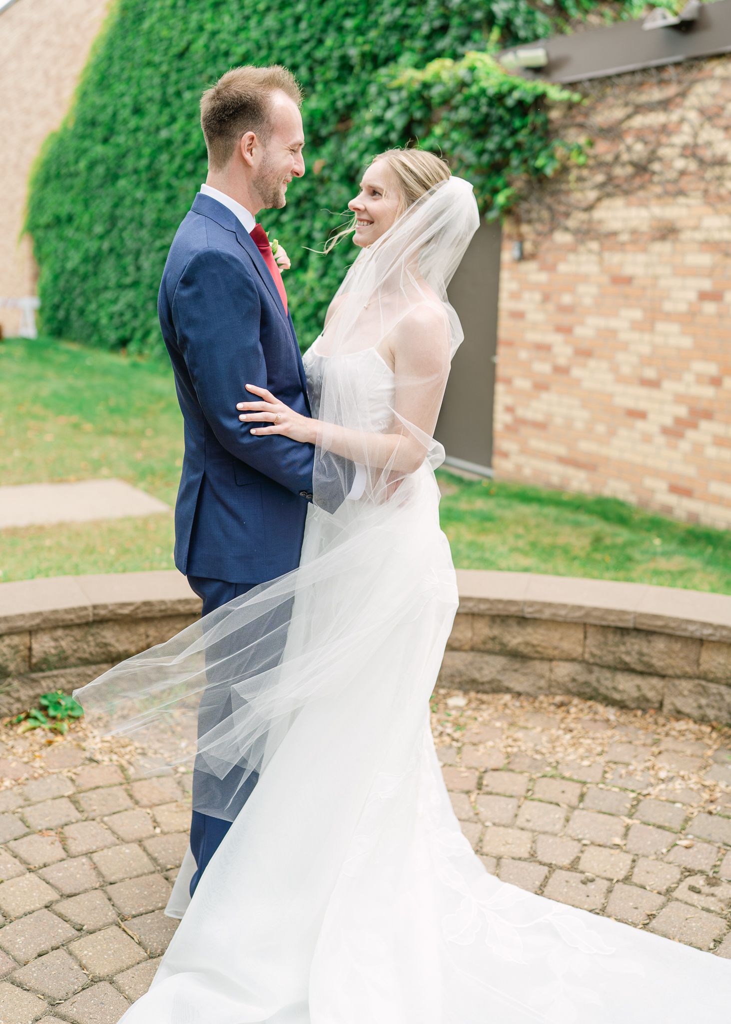 Bride in a white gown and long veil embraces groom in a navy suit and red tie on a brick patio near a vine-covered wall.