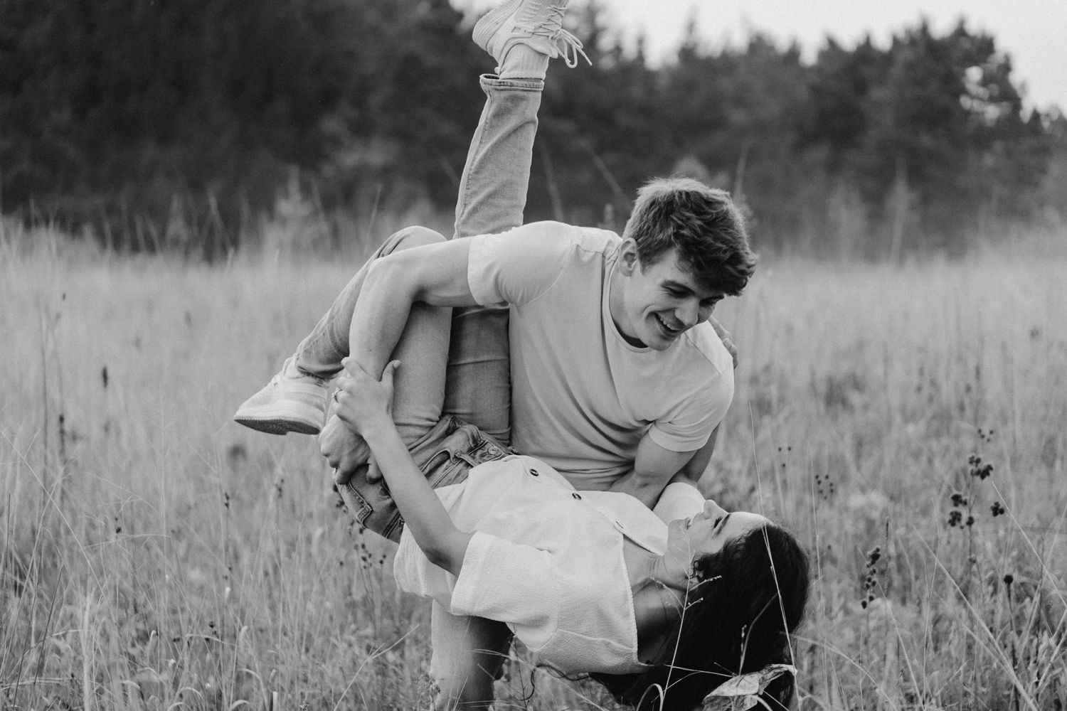 Black and white photo of a playful couple laughing as a man lifts a woman in a tall grassy meadow with trees behind them.