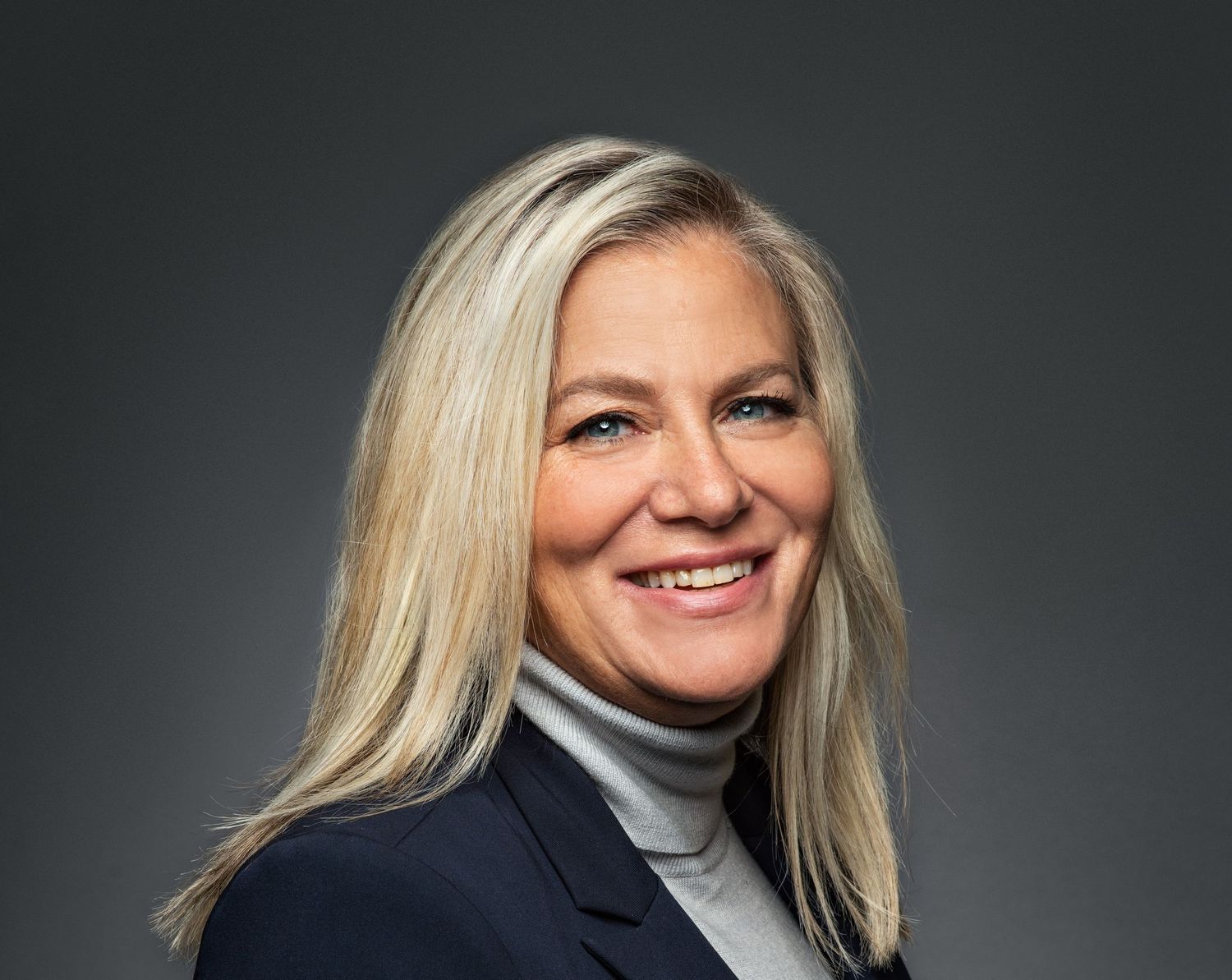 Professional headshot of a smiling blonde woman in a navy blazer and white turtleneck against a gray background.