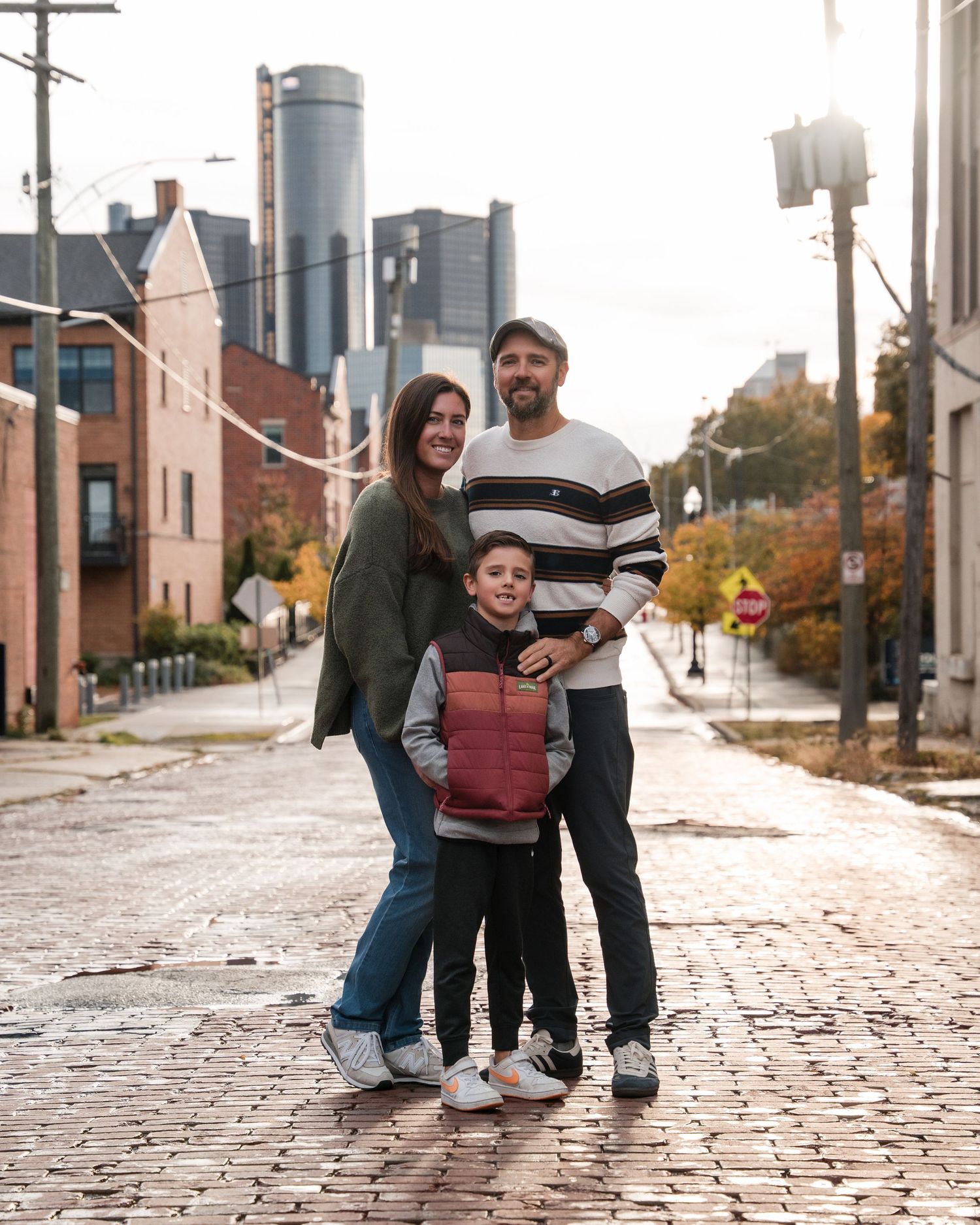 Family of three posing on a cobblestone street in Detroit with the Renaissance Center skyline in the background.