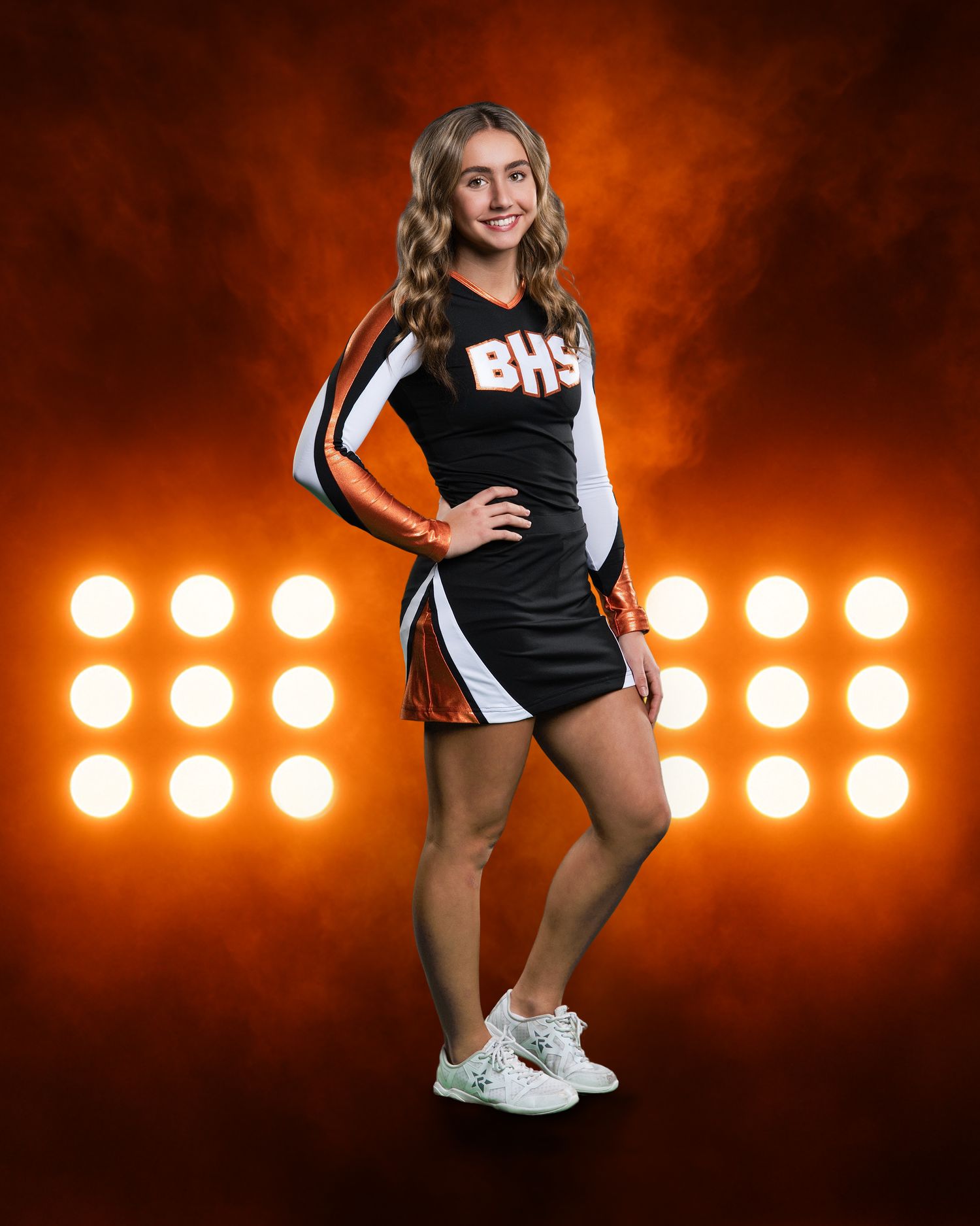 BHS cheerleader in black and orange uniform poses confidently against a glowing orange stadium lights backdrop.