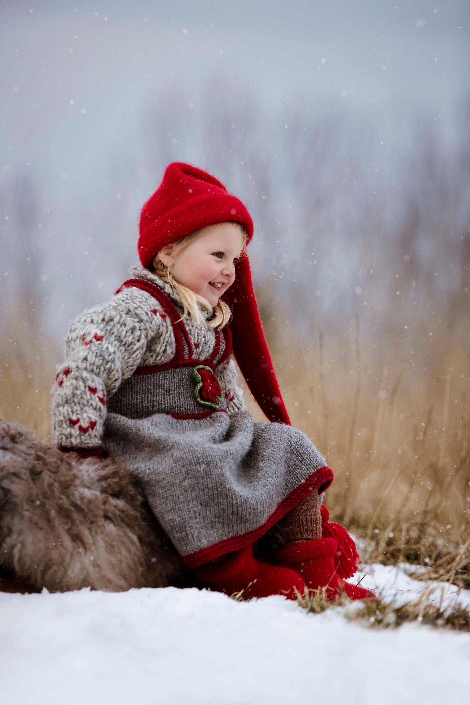 Winter scene with small figure in red hat and gray sweater sitting among dry grass and snow.