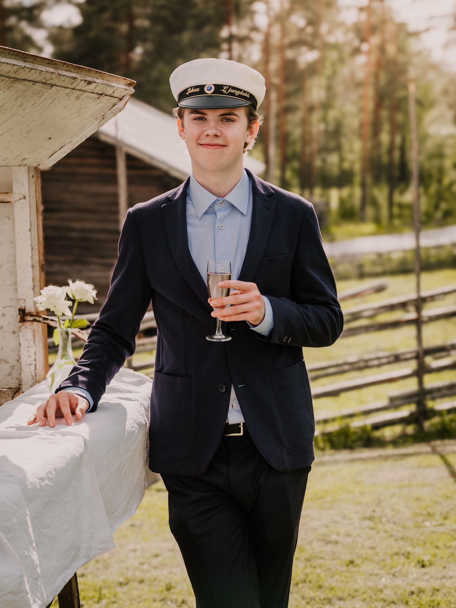 Young man in navy suit and captain's hat holding champagne flute at outdoor celebration.