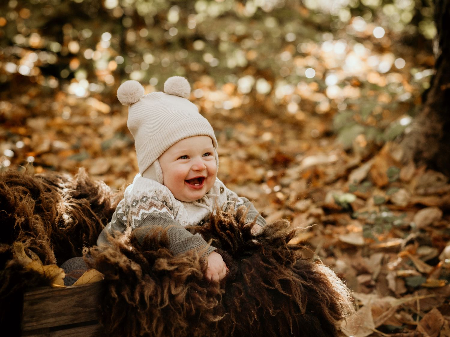 A joyful baby wearing a white winter hat laughs while lying on autumn leaves.