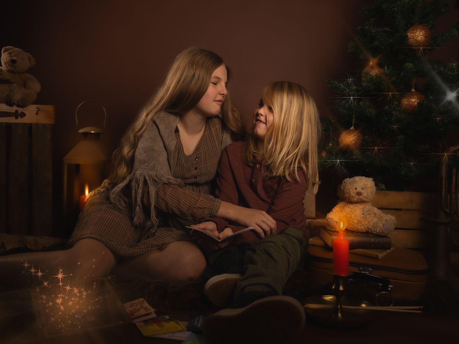 Two girls share a warm moment by a Christmas tree with candles and teddy bears in a cozy holiday setting.