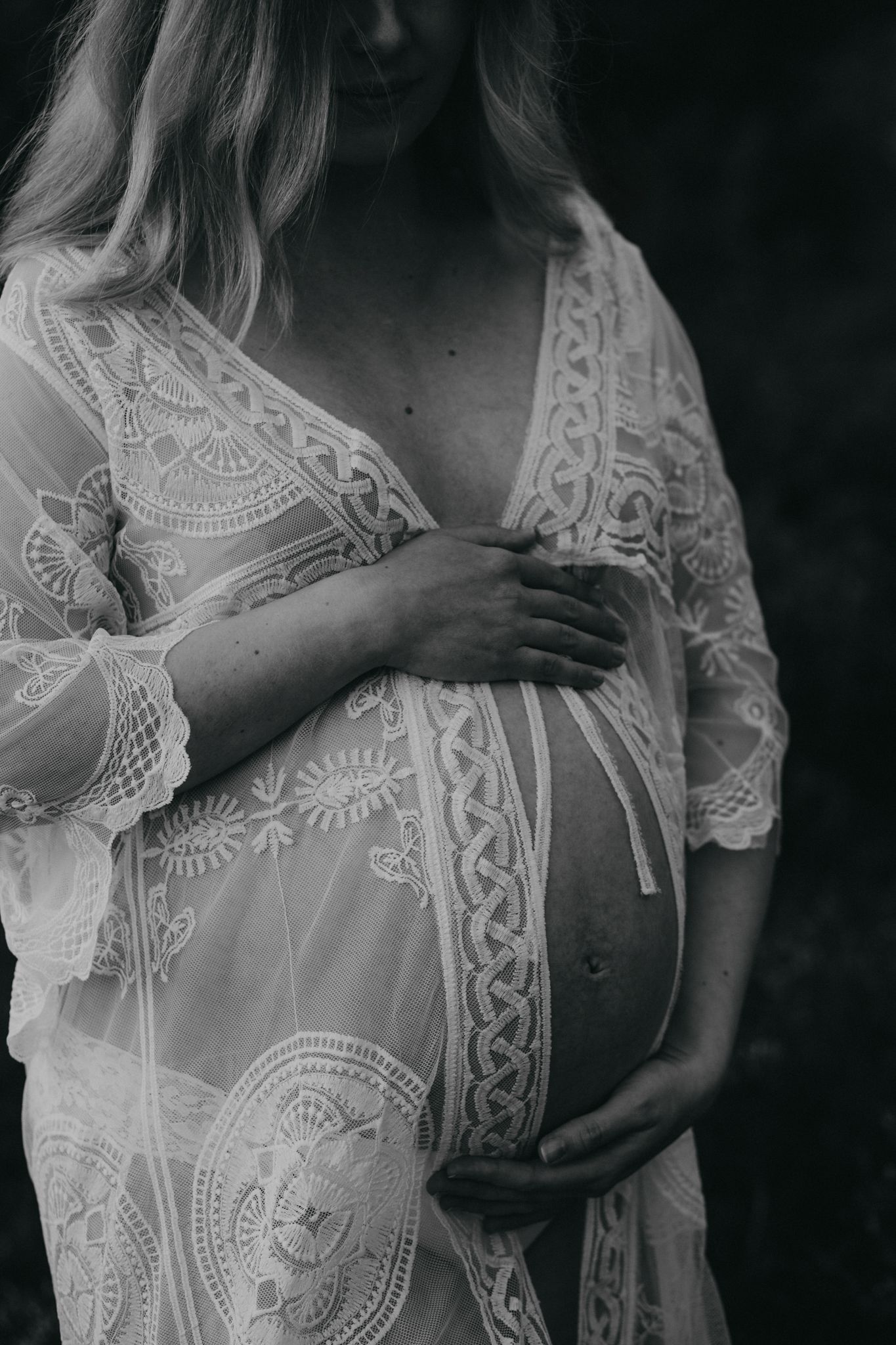 A black and white maternity photo showing a pregnant belly draped in delicate white lace and embroidered fabric.