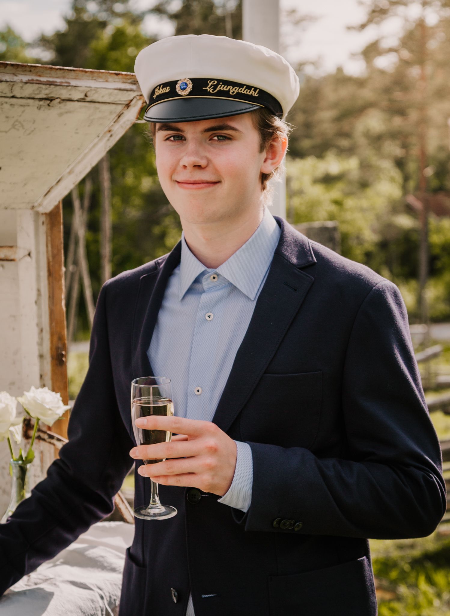 Young man in navy suit and white captain hat holding champagne flute outdoors at a formal celebration.