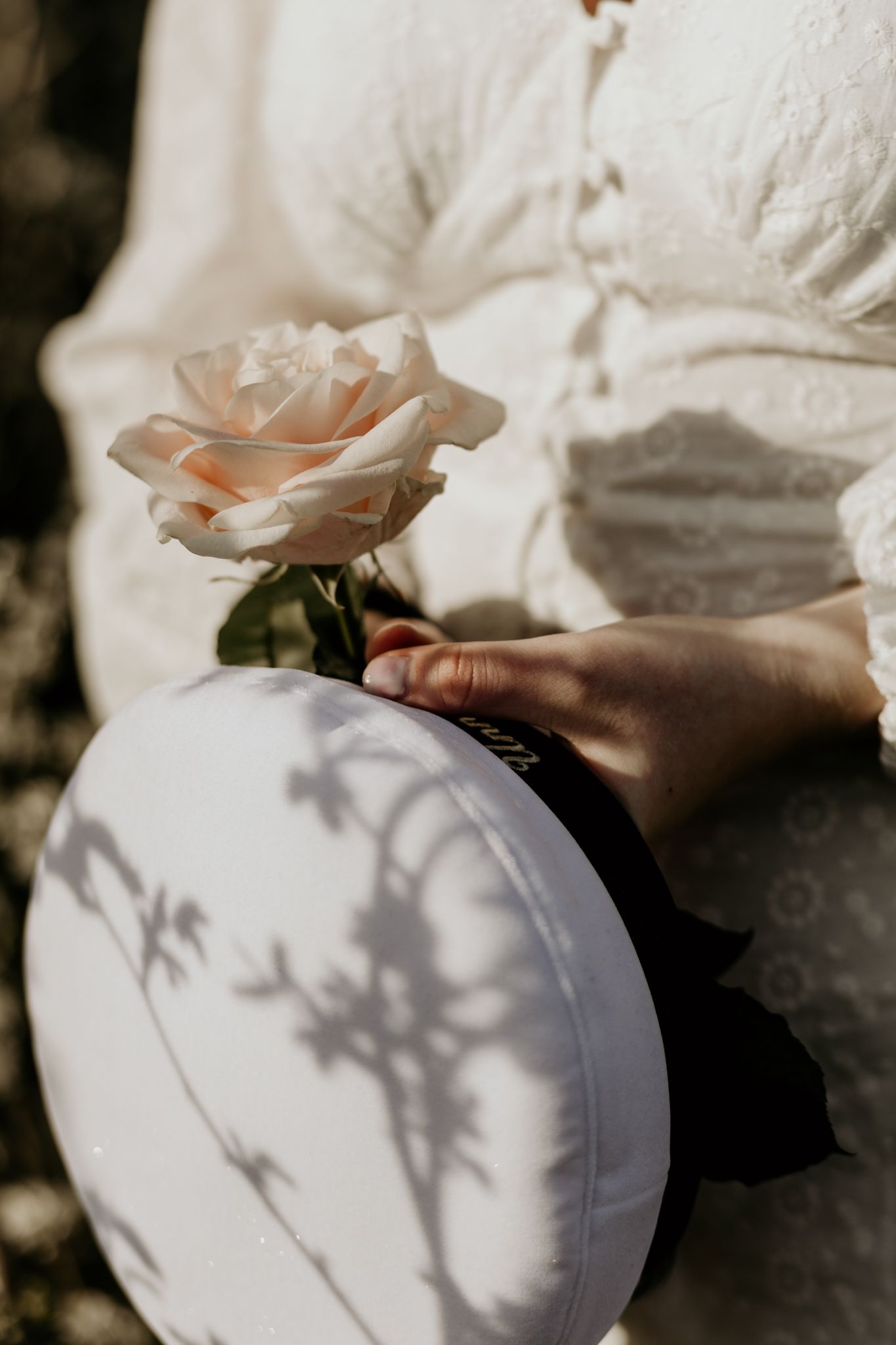 Bride in white lace dress holding a blush pink rose and round white clutch with floral shadows in sunlight.