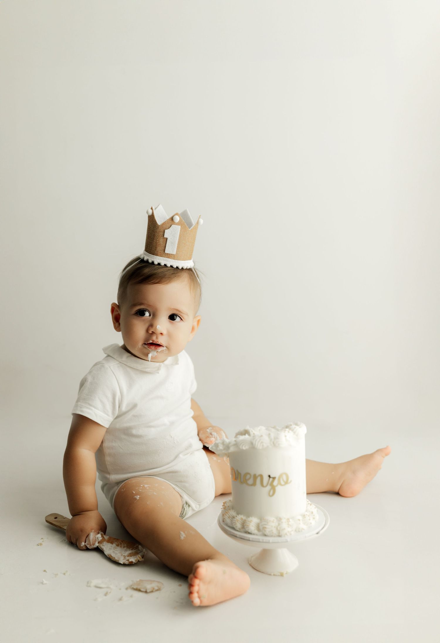 Baby in white onesie wears gold crown while sitting next to small white cake.