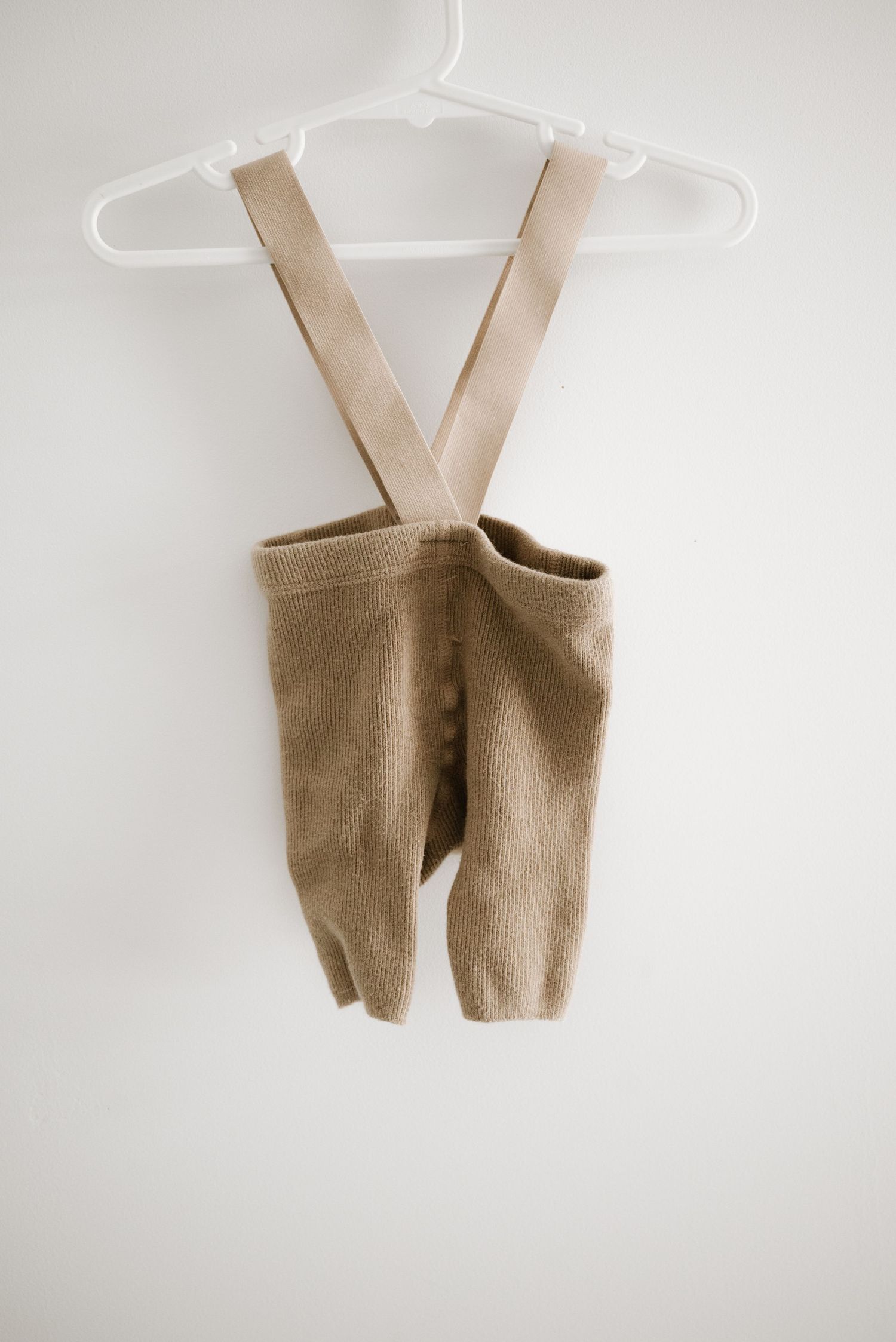Tan knit baby romper with suspender straps hanging on white hanger.