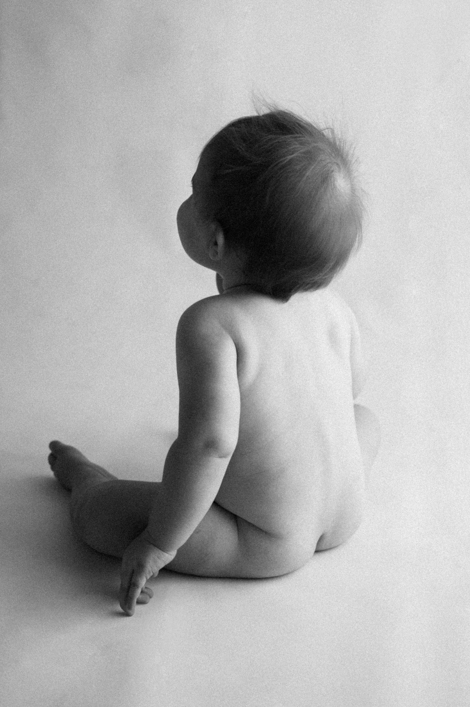 Black and white artistic portrait of a baby sitting facing away from camera against white background.