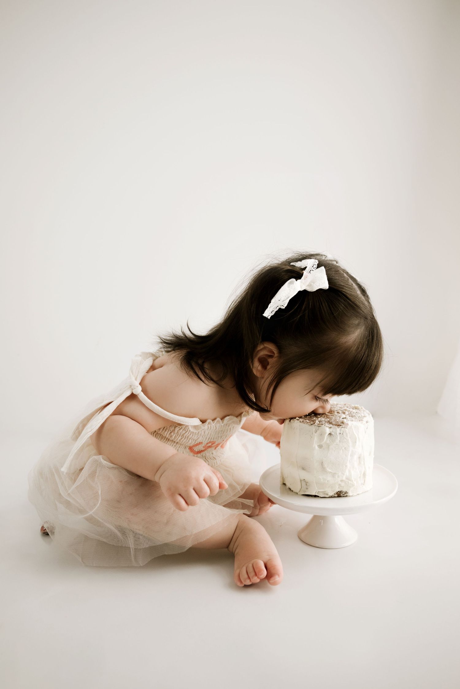 Toddler in a white tulle dress sits next to a small white cake on a pedestal against a light background.