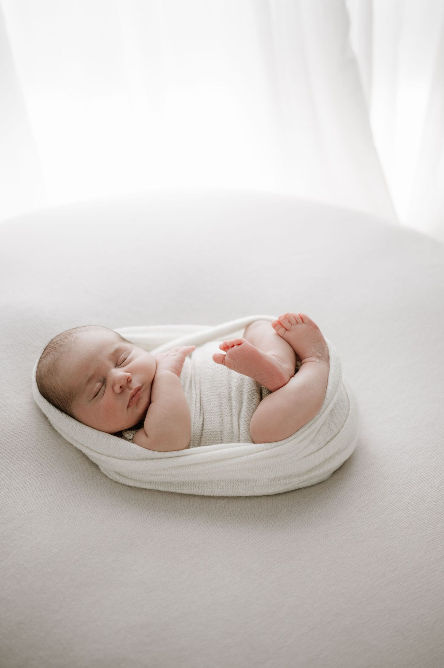 A newborn baby swaddled in white fabric sleeps peacefully in a soft, curved cushion.