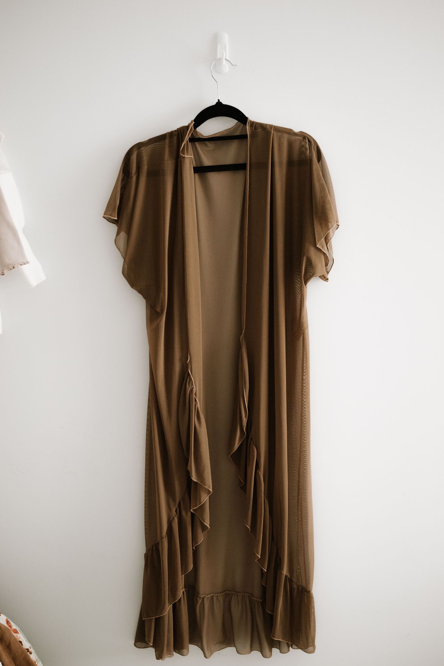 Long brown flowing maxi cardigan with ruffled hem hanging on white plastic hanger against neutral wall.