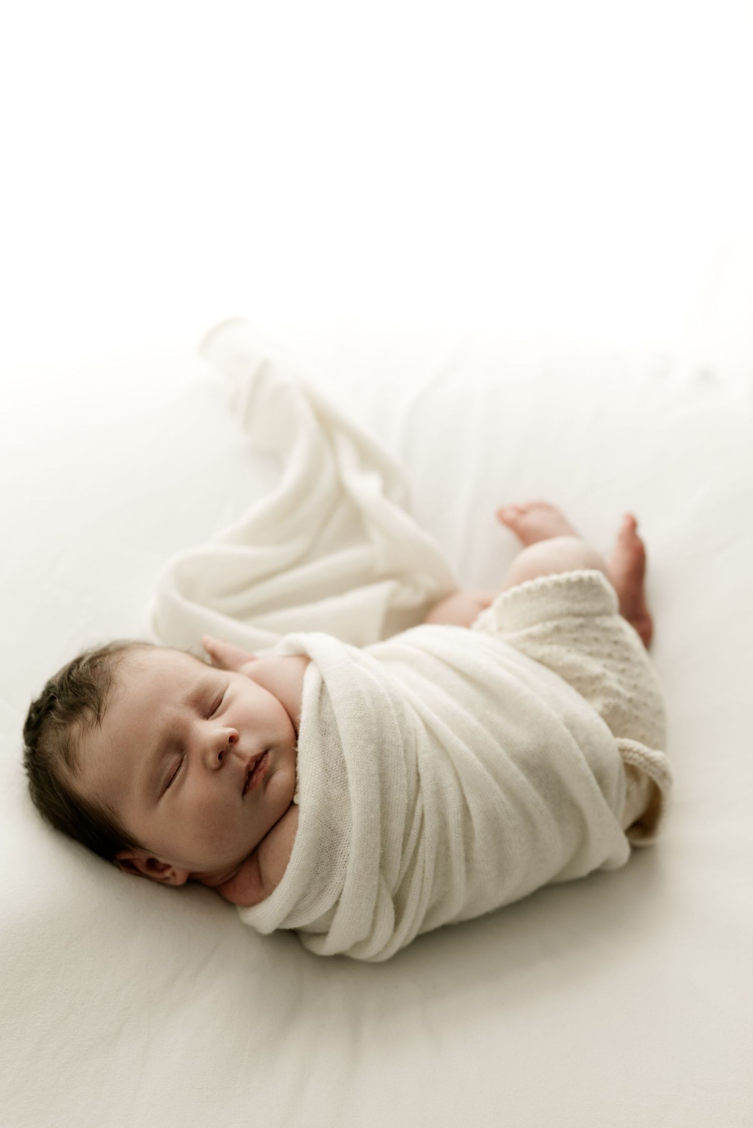 Sleeping newborn wrapped in a soft white blanket on a white background.