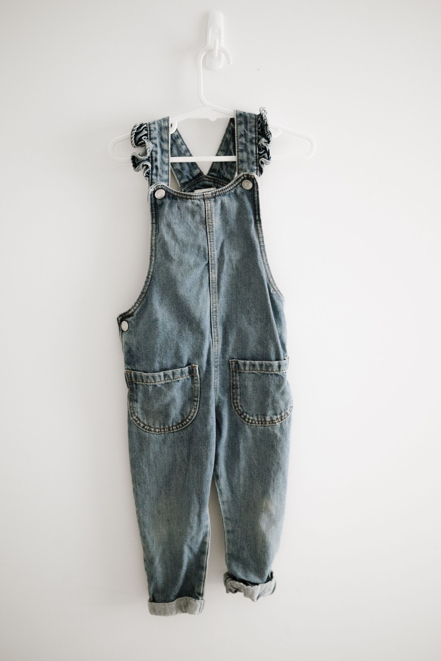 Vintage-style denim overalls with ruffle straps and front pockets hanging on white hanger.