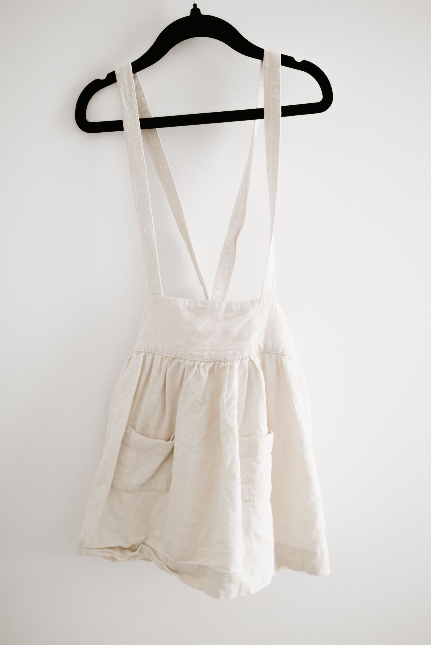 White cotton pinafore dress with crossback straps and side pockets hanging on black hanger against white wall.