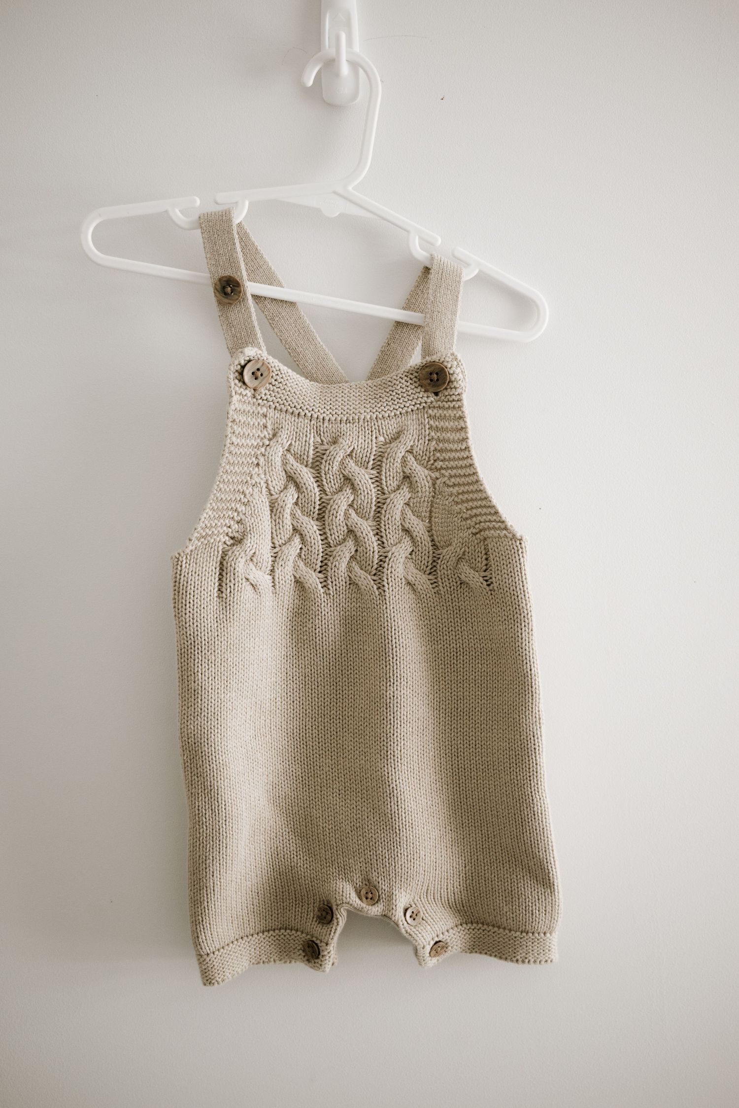 Beige knitted baby romper with cable knit pattern and shoulder straps on hanger.