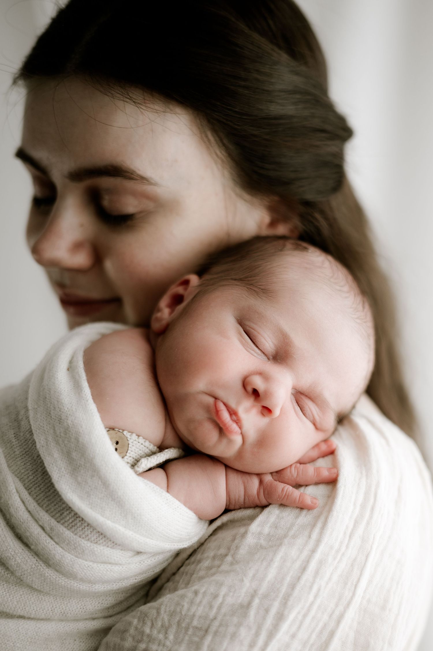 A sleeping newborn wrapped in white fabric is held close while being tenderly embraced.
