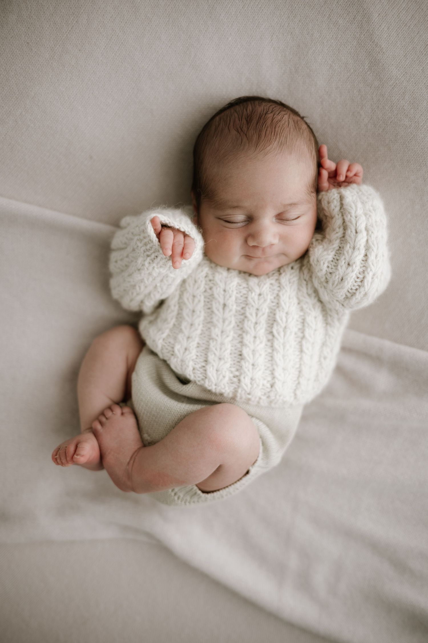 Sleeping newborn wearing cozy white knit sweater lying on soft cream fabric with tiny feet showing.