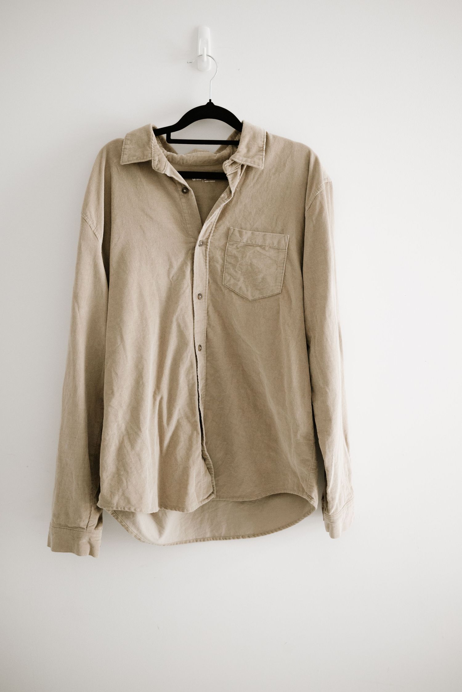 A beige button-down shirt with a chest pocket hanging on a white wall.
