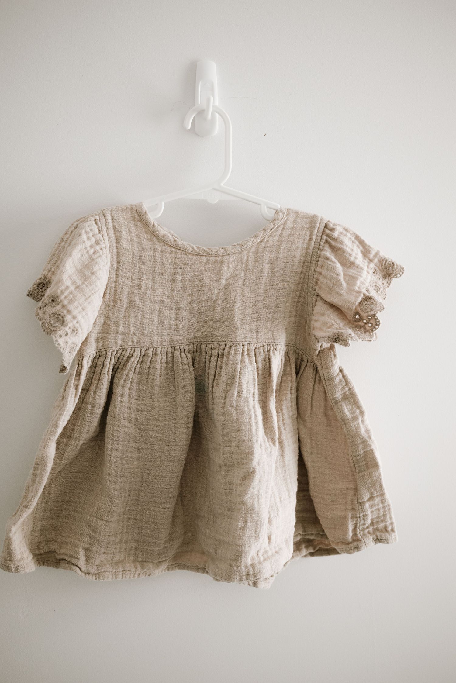 A beige linen baby dress with puff sleeves and gathered bodice hanging on a white wall.