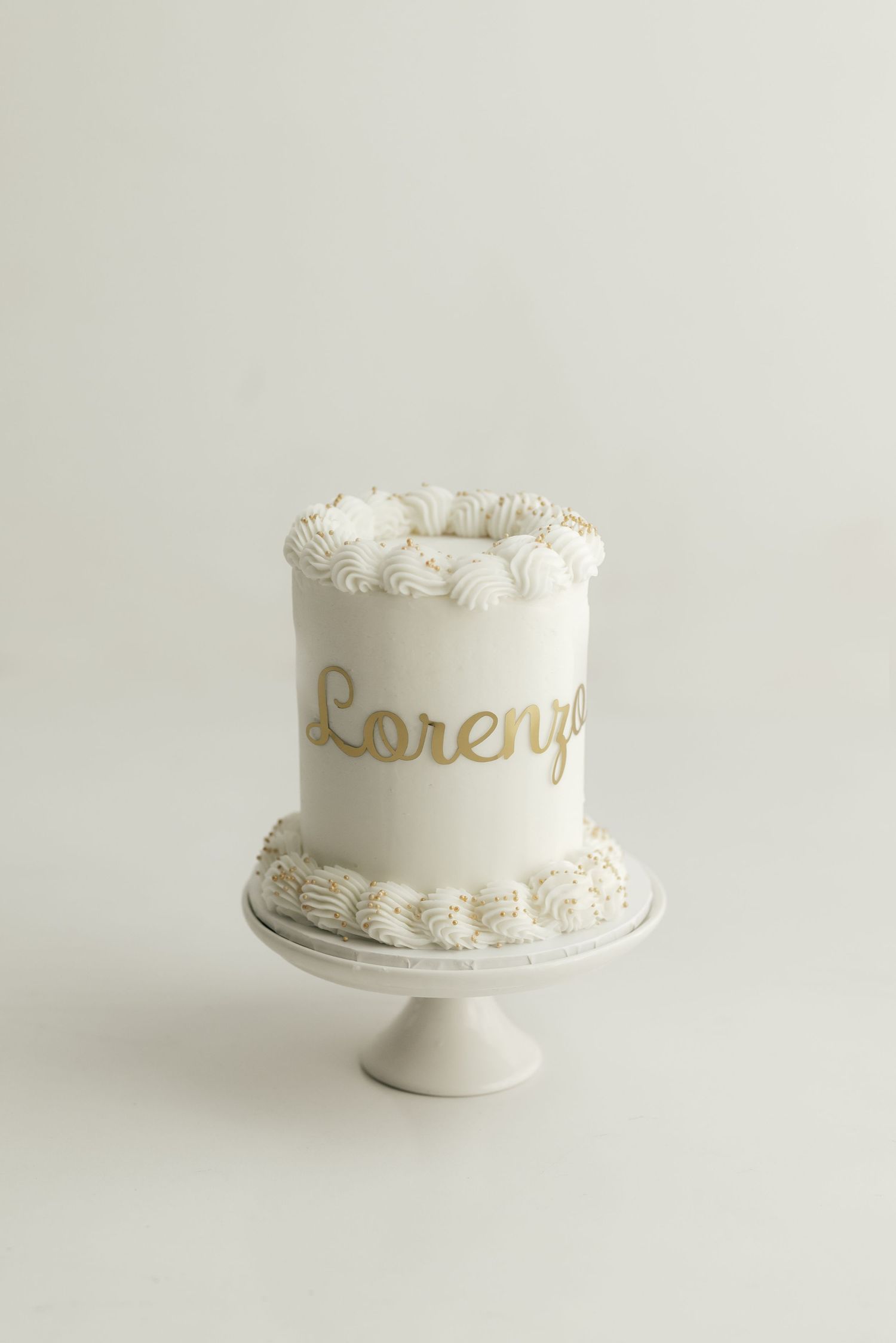 A white birthday cake with gold lettering and piped frosting on a cake stand.