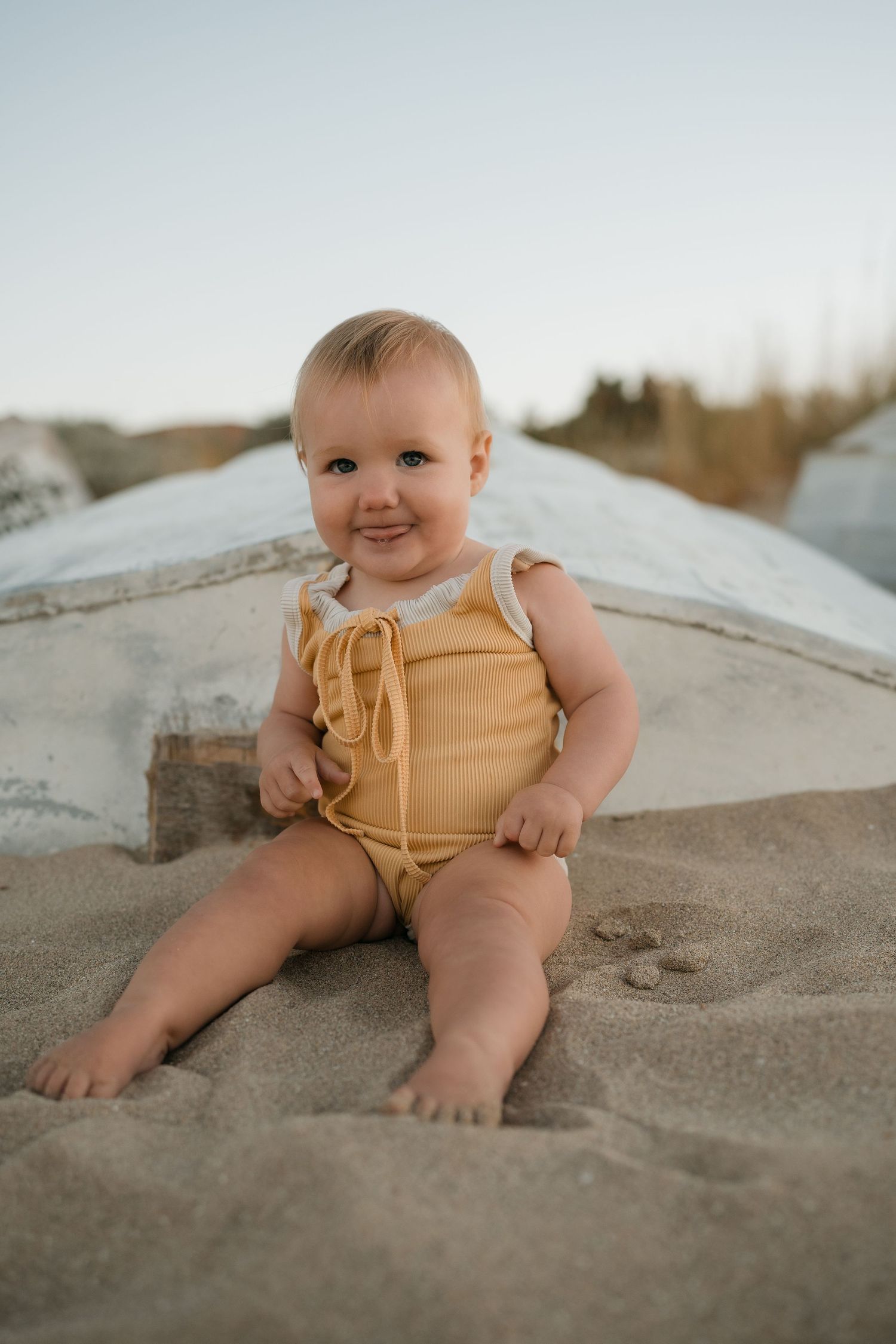 Baby in yellow romper sitting on sandy beach during sunset photoshoot.