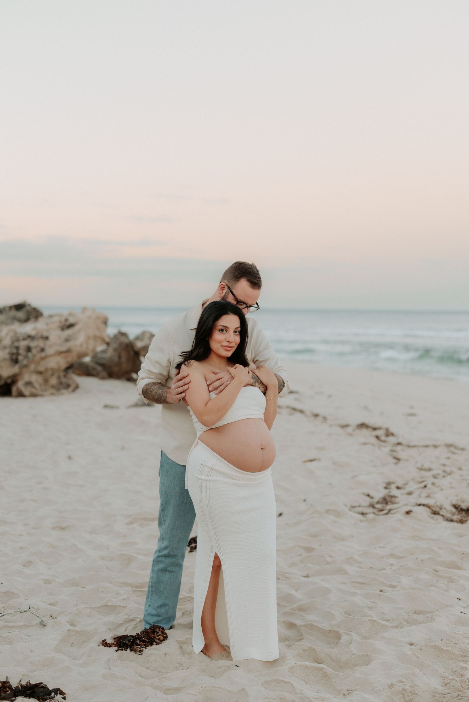 A romantic maternity photoshoot on a beach at sunset shows expectant parents embracing on the sand near rocks and ocean waves.