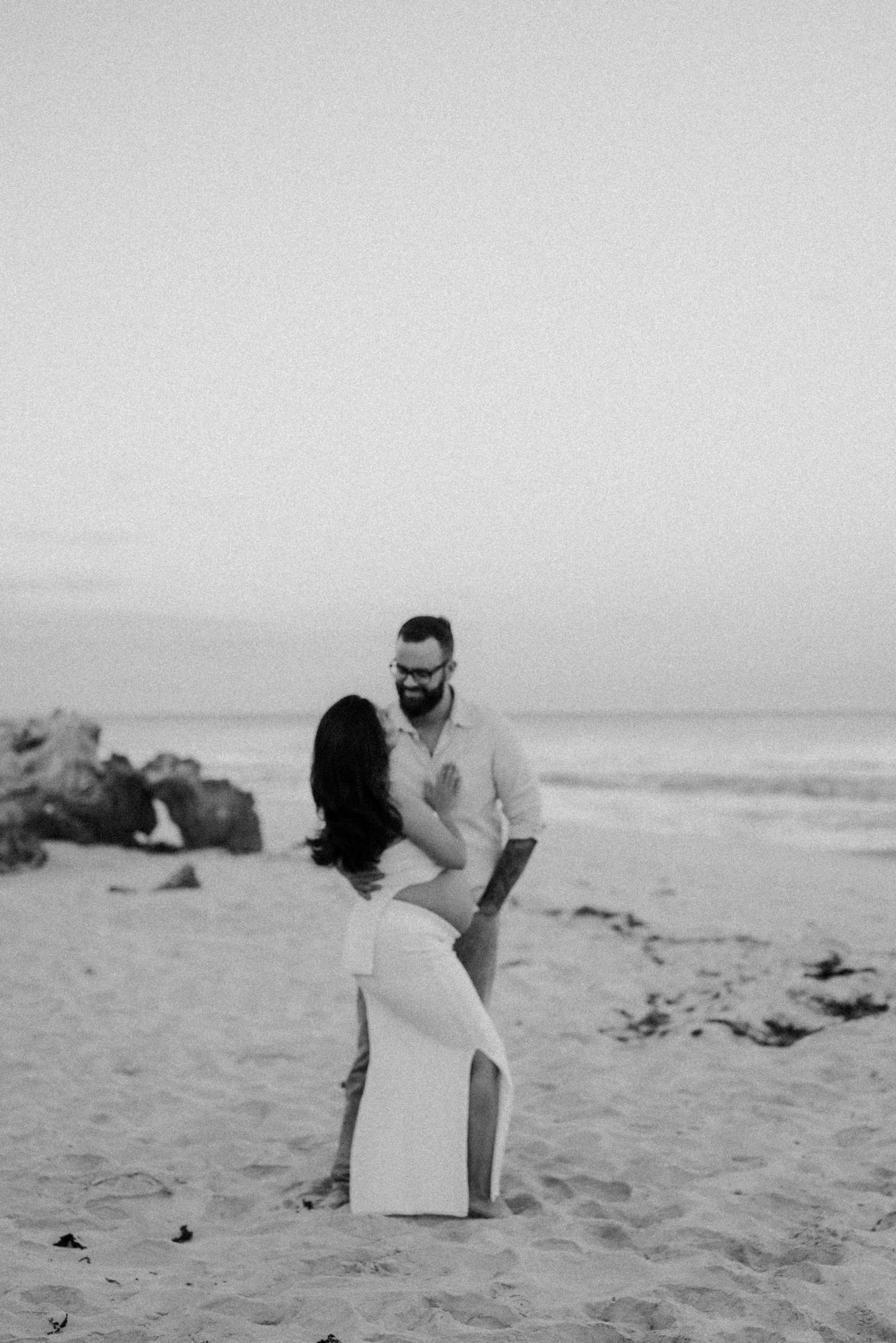 Black and white beach portrait of couple in romantic pose near ocean rocks.