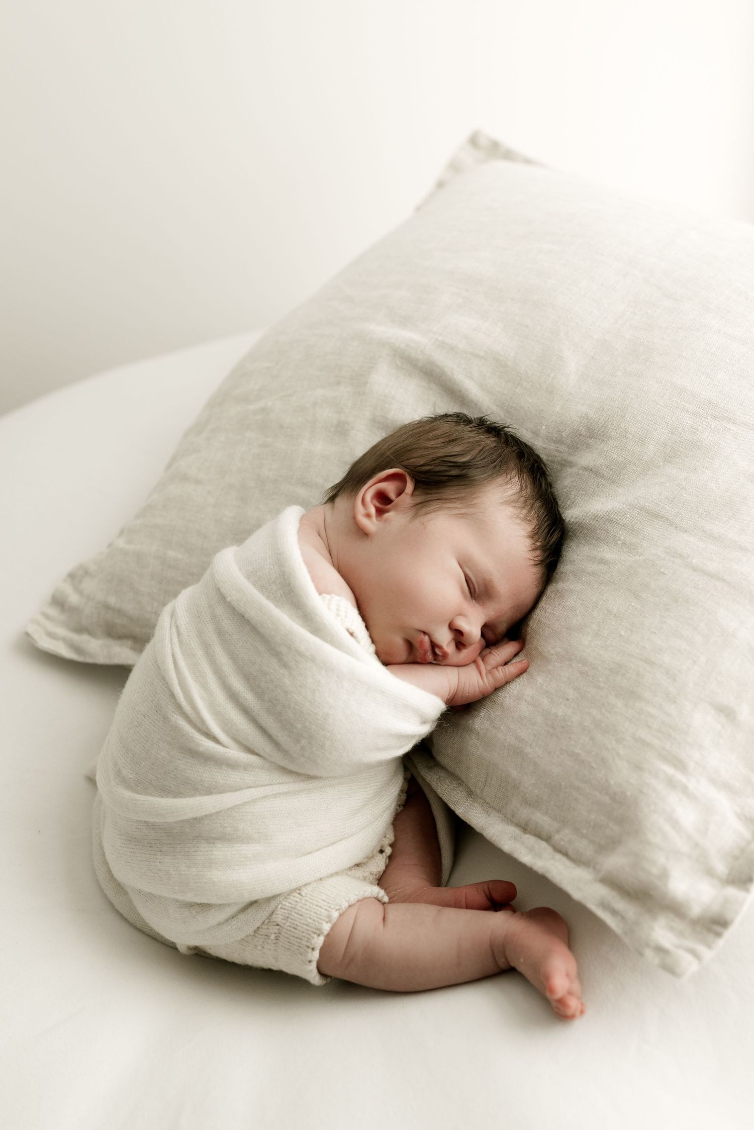 Sleeping newborn swaddled in white wrap resting on light gray pillow.