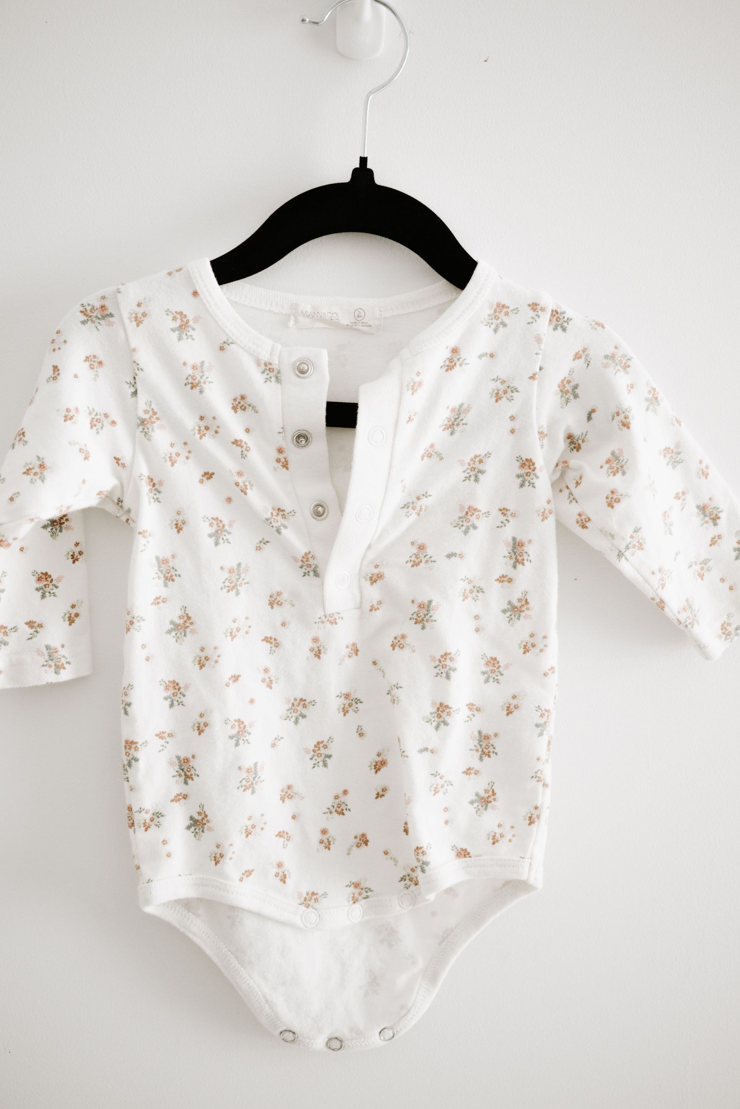 White baby onesie with delicate floral print and henley neckline hanging on black hanger.