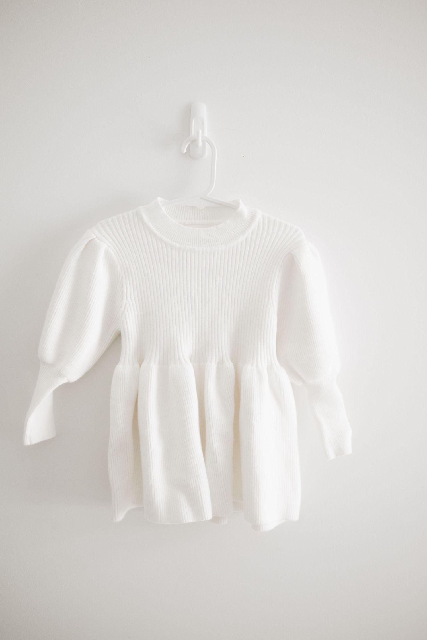 A white ribbed knit baby dress with three-quarter length sleeves hanging on a white wall.