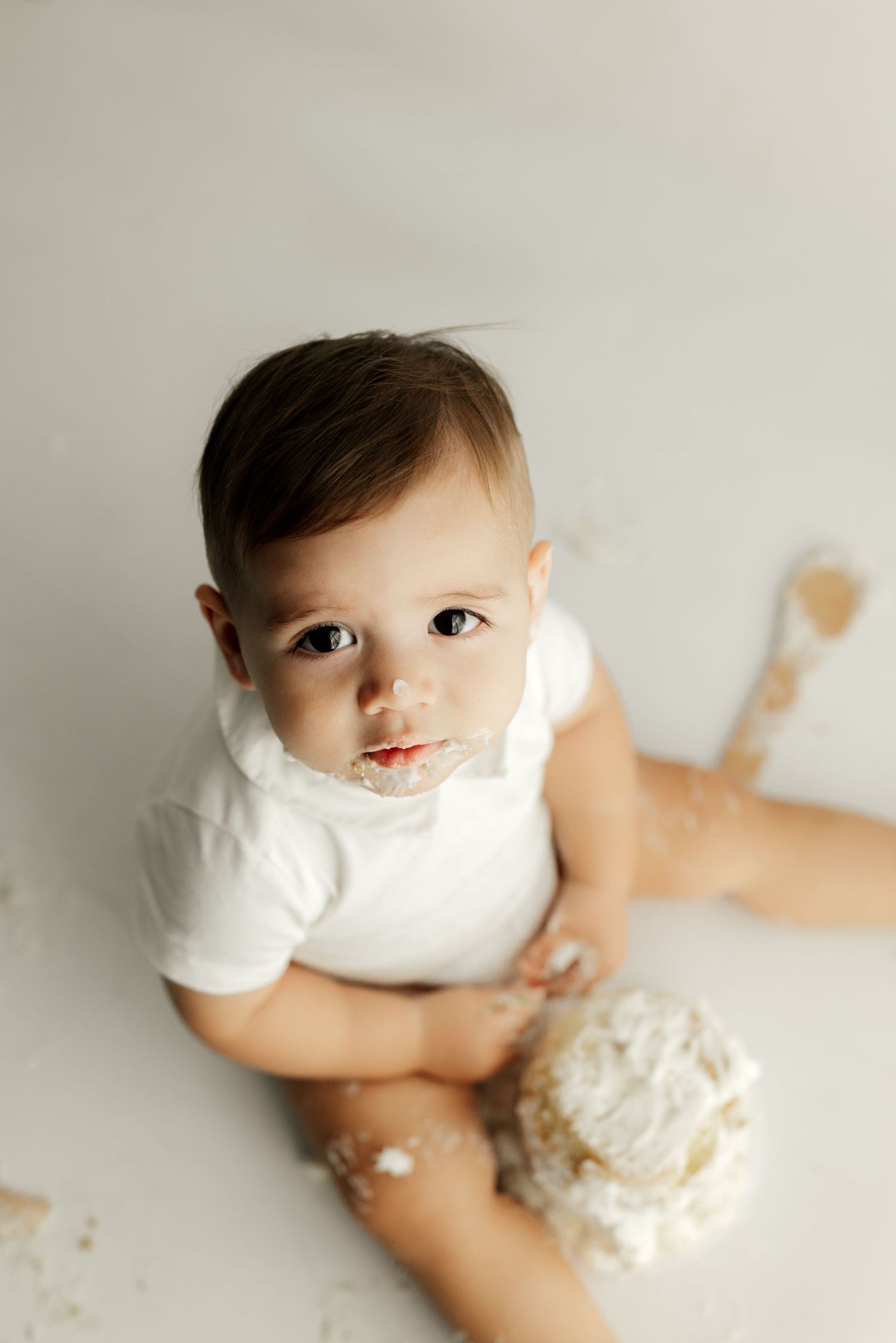 A baby in white clothing sits on the floor with cake smeared around their mouth.