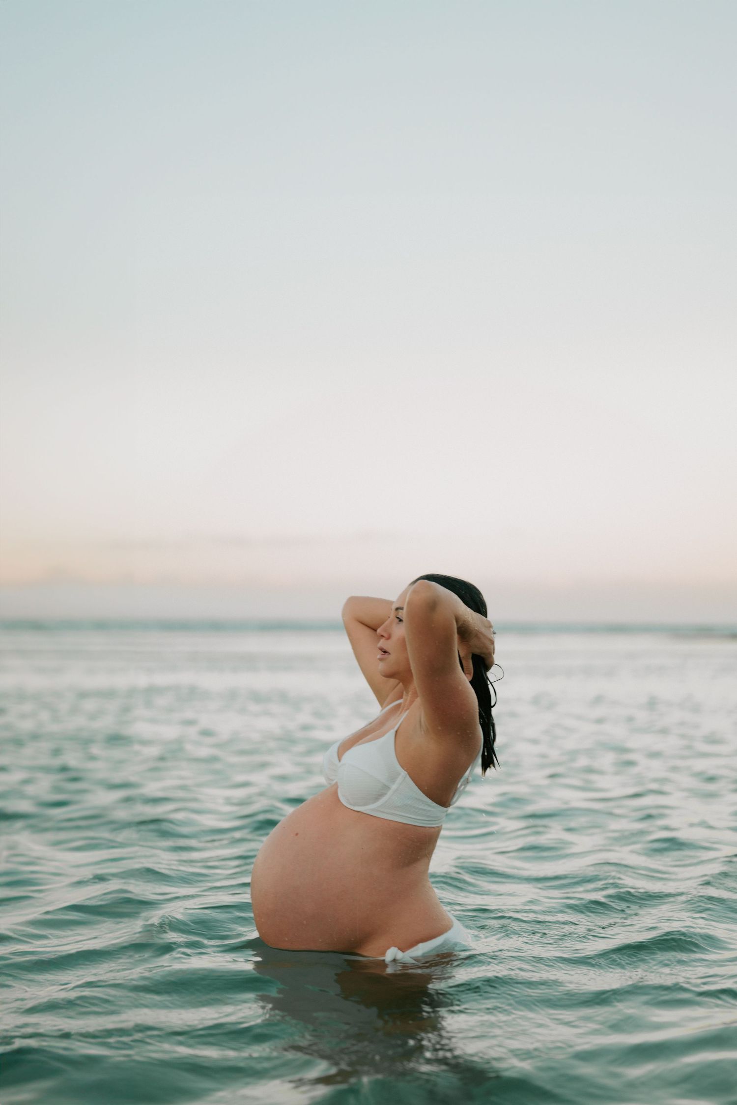 Pregnant woman in white bikini standing in ocean at sunset.