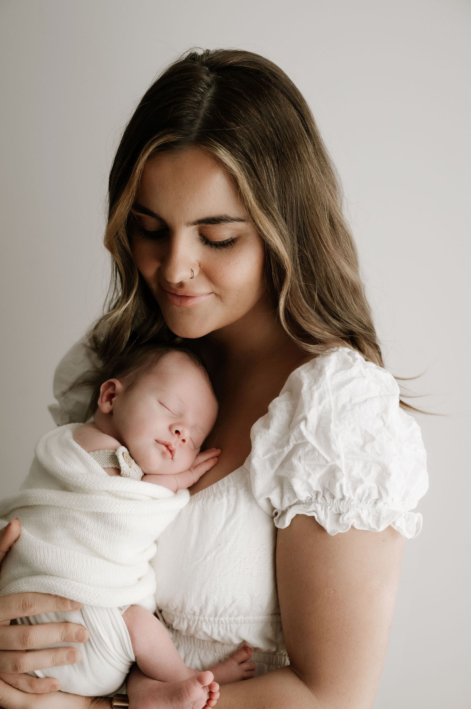 Mother in white dress holds sleeping newborn wrapped in white blanket against neutral background.