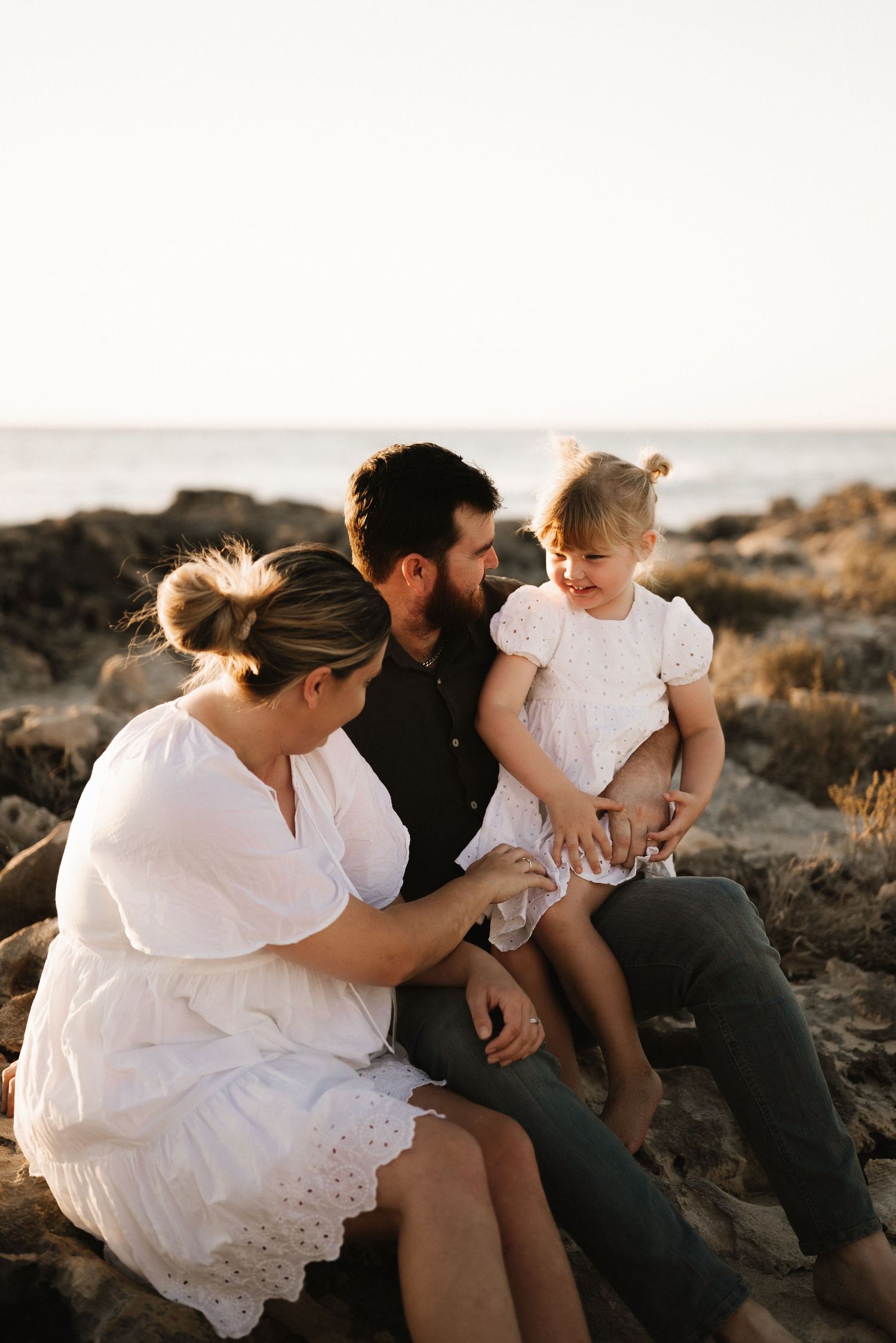Family sits together on rocky coastline during golden hour photo session.