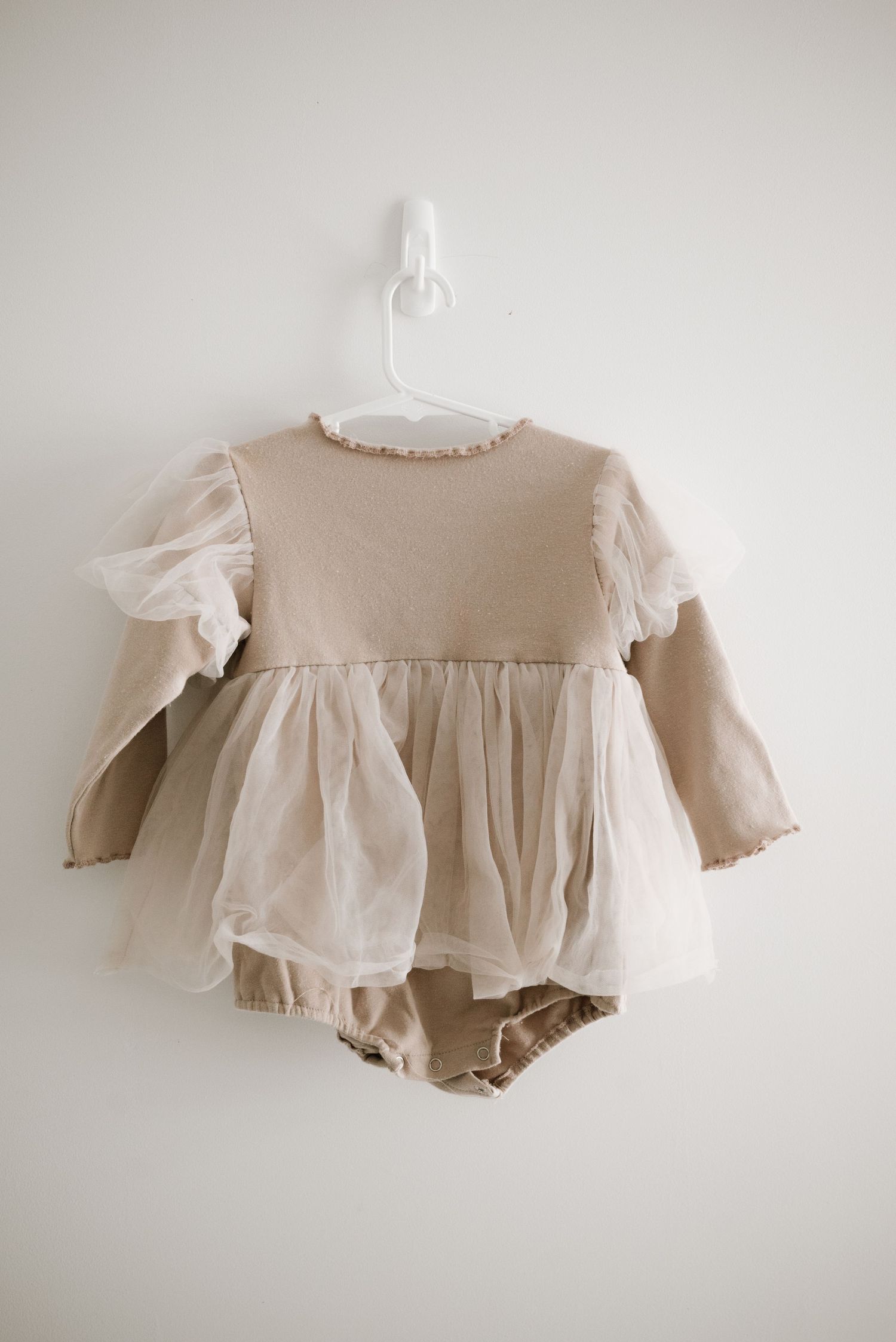 Beige baby dress with white puff sleeves and matching bloomers hanging on white hanger against neutral wall.