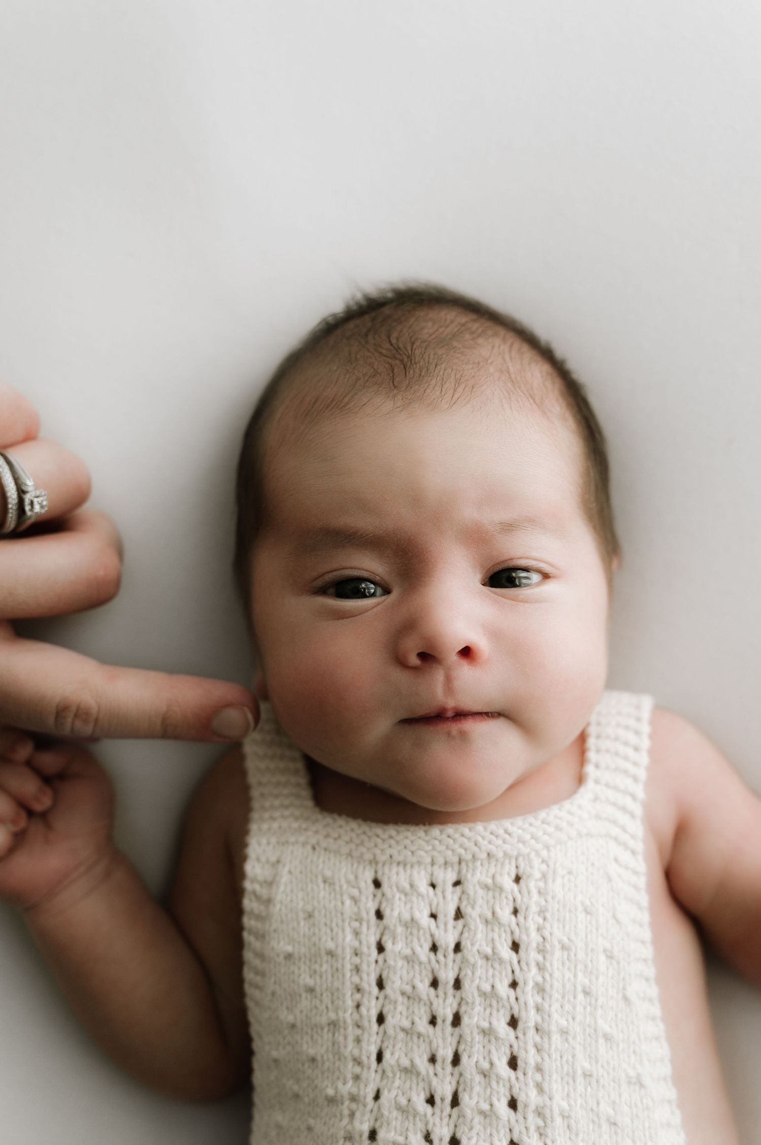 A baby wearing a white knit sleeveless top lays on a light background while someone's hand touches their cheek.